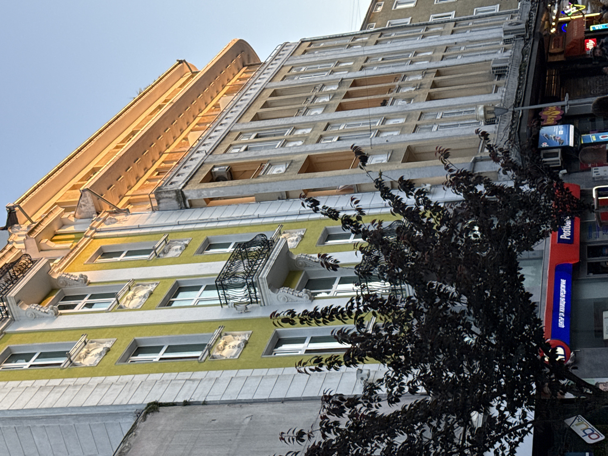 The image shows a street view of buildings with distinct architectural styles under a clear blue sky. The building on the left is painted in a vibrant lime green and white, featuring ornate decorative elements above some of the windows, including relief sculptures. A black wrought-iron balcony juts out from this building. To its right, a taller, more modern building stands, characterized by a grid-like facade with vertical columns and rows of evenly spaced windows. The top floors of this building are bathed in warm, golden light, suggesting it's late afternoon or early morning. In the foreground, a dark-leaved tree partially obscures the lower portions of the buildings, with its branches reaching upwards. Several storefronts are visible at street level, including one with a red and blue sign that reads "ЦЕНТЪР ЗА ЖИЛИЩНО КРЕДИТИРАНЕ" and "Postbank". Other signs are partially visible, suggesting a commercial area. The overall scene depicts an urban environment with a mix of older and newer architectural elements, illuminated by natural light.