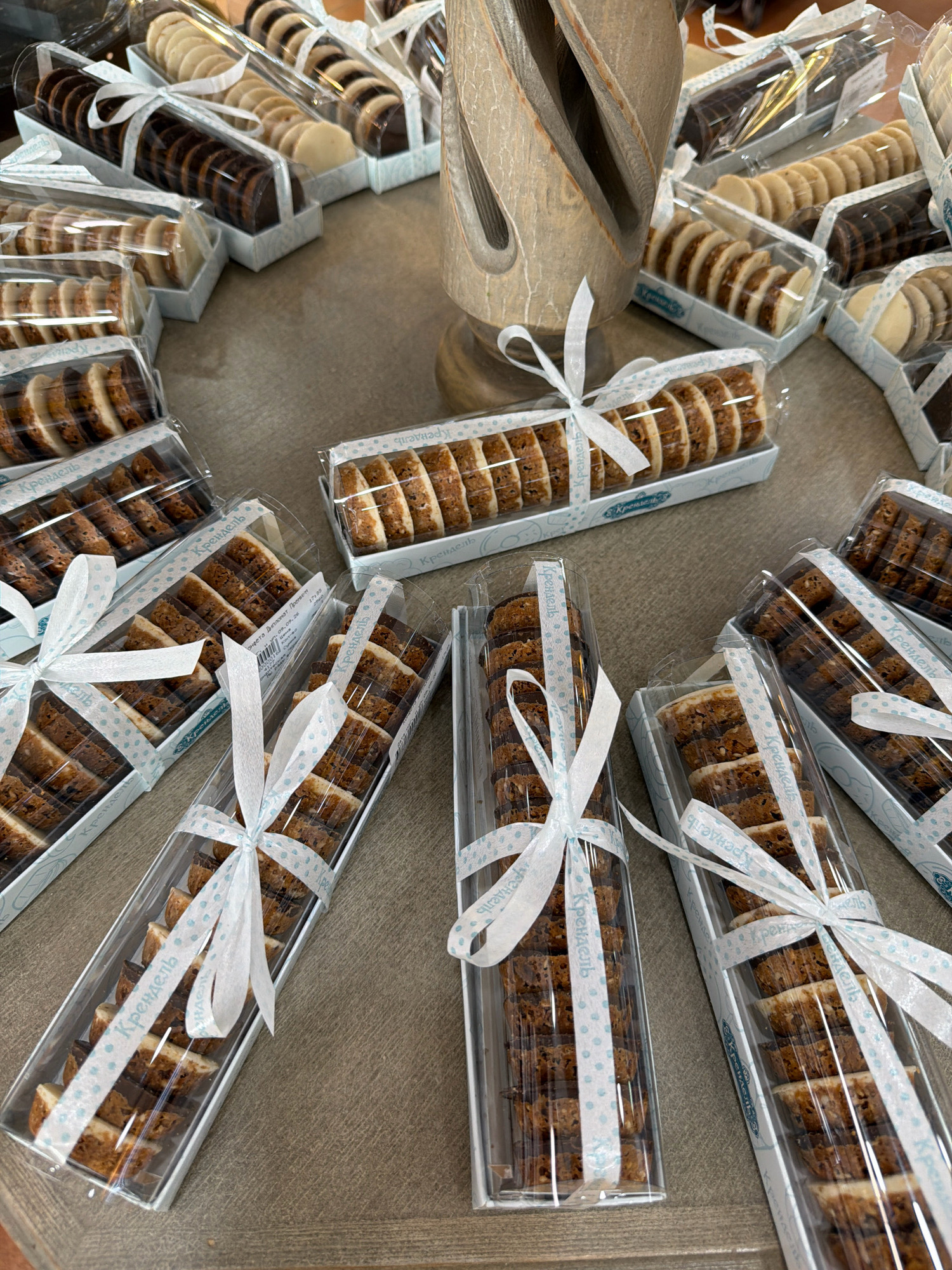 The image depicts an arrangement of assorted cookies packaged in clear plastic boxes, each tied with a white ribbon. The cookies appear to be layered, with some boxes containing round cookies of alternating dark and light colors, and others containing rectangular cookies with a striated pattern. The boxes are placed on a textured surface that resembles a wooden table. In the background, a portion of a decorative wooden sculpture is visible. There are no people or active interactions depicted in the scene. The overall impression is one of a retail display or a prepared product offering. The location in Pavlodar, Kazakhstan cannot be determined from the provided image.