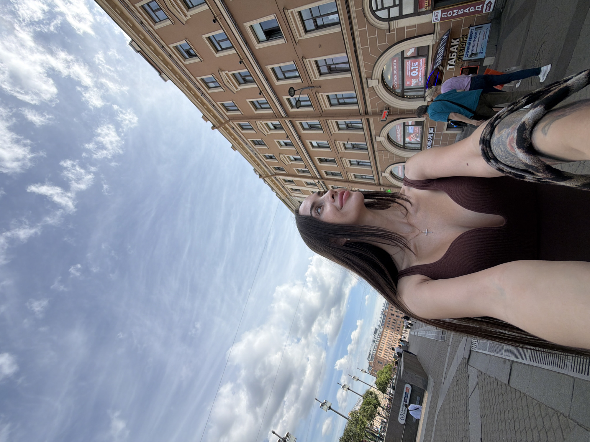A woman with long dark hair and a brown top is taking a selfie outdoors. She is looking up and to the side, with a slight smile. A silver cross necklace is visible on her chest. The background shows a classic European-style building with multiple windows and arched storefronts. One storefront has Cyrillic text advertising "ТАБАК" (tobacco) and "VAPE". Another sign advertises "ЛОМБАРД" (pawnshop) and "СКУПКА ЗОЛОТА" (gold buying). Several other people are walking on the street in the background, and there are streetlights and trees. The sky is blue with scattered white clouds. The image appears to be taken during the daytime. The overall scene is of a bustling city street in Saint Petersburg, Russia.