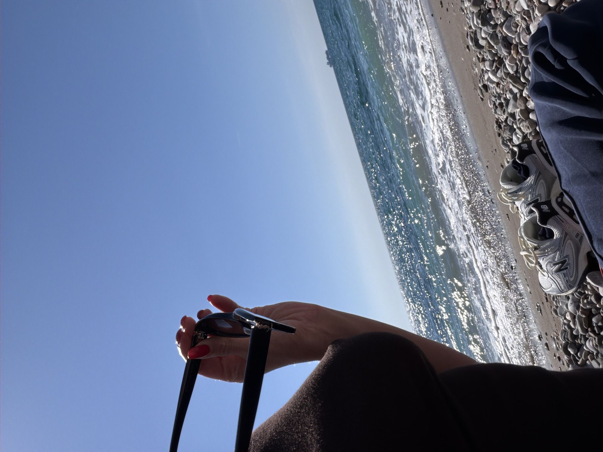 A first-person perspective shot depicts a woman sitting on a pebble beach in Bar, Montenegro. In her left hand, she holds a pair of sunglasses. Her fingernails are painted bright red. The background shows the Adriatic Sea, with sunlight glinting off the waves. To her right, a pair of silver New Balance sneakers rests on the pebble beach, next to a dark blue garment. The sky above is a clear blue.