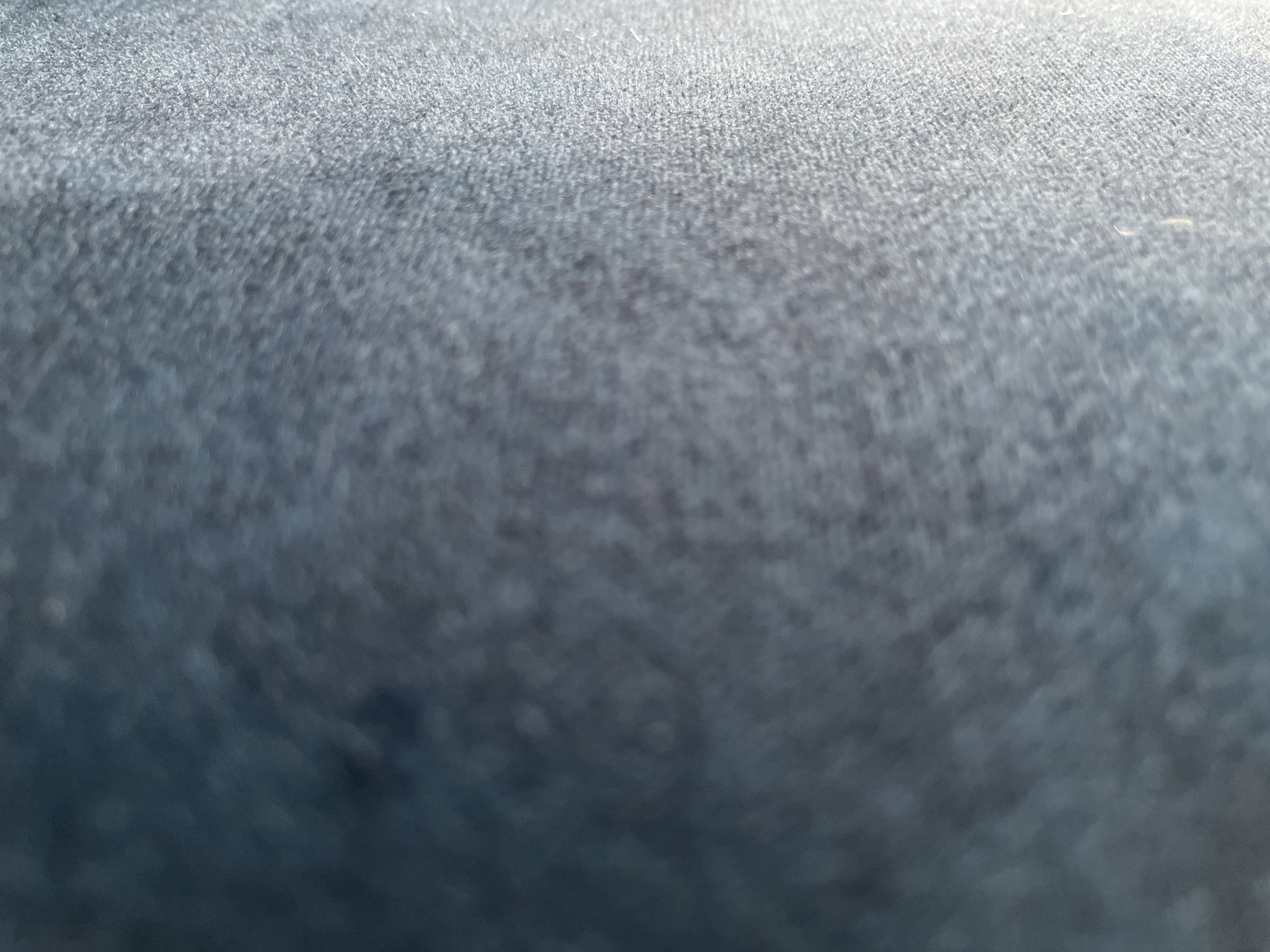 The image is a close-up, abstract shot of a dark blue, textured fabric. The focus is shallow, with the upper portion of the image appearing sharper than the lower portion, which is blurred. The lighting creates a gradient, with a bright highlight in the upper right corner that fades into darker tones towards the bottom. The texture of the fabric appears to be woven, with a slightly fuzzy or fibrous surface. No specific location, people, or objects are identifiable within the frame.