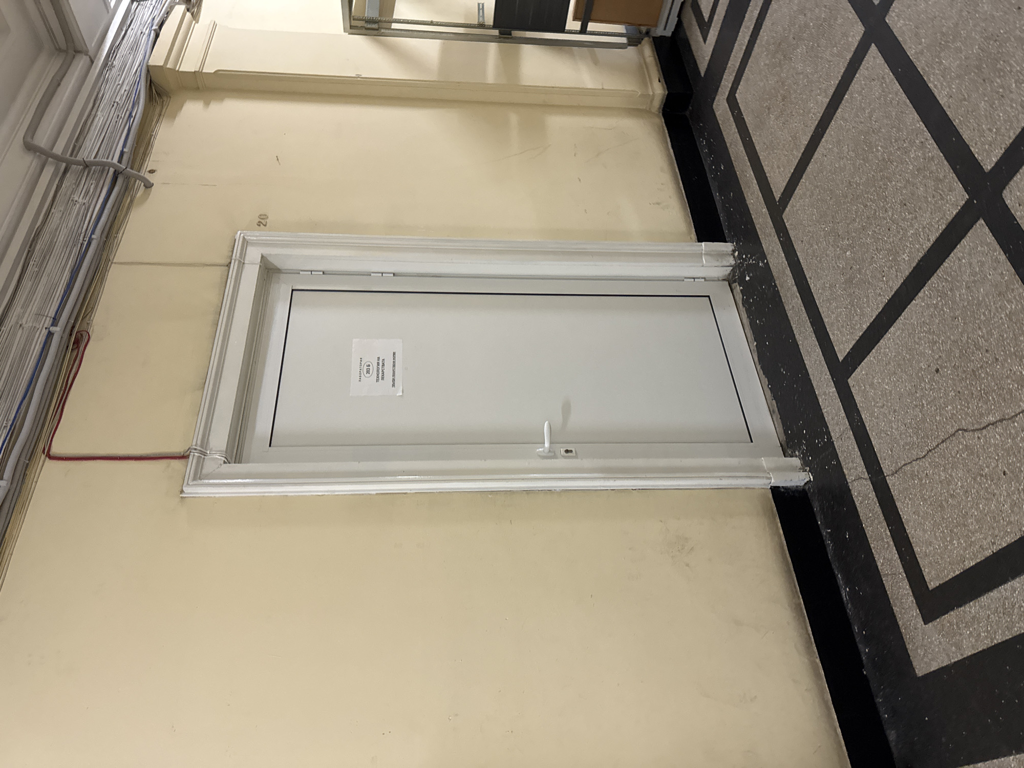 The image shows a white door in a beige wall, with a white frame. The number "20" is faintly visible above the door frame. There is a document affixed to the door, and a small white handle. To the left of the door, electrical wiring is visible running along the wall. To the right, the edge of a tiled floor with a geometric pattern can be seen. The specific location in Sofia, Bulgaria cannot be determined from the image alone.