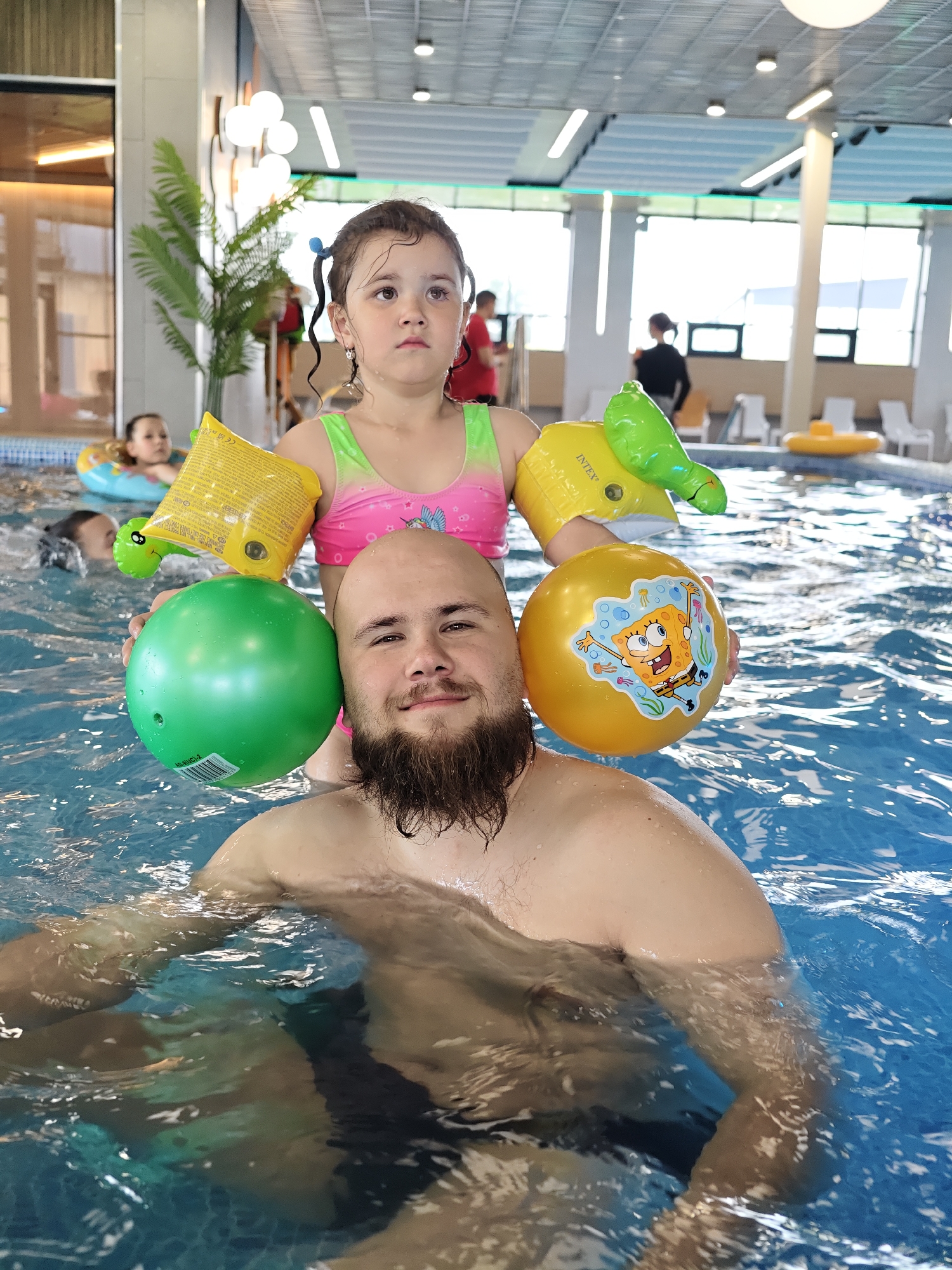 The image captures a scene inside what appears to be an indoor swimming pool, likely in Votkinsk, Russia. A man with a beard, bald head, and fair complexion stands in the water, submerged to his chest. He is holding two inflatable balls, a green one on his left side and a yellow one with a SpongeBob SquarePants design on his right.
Perched on his shoulders is a young girl with pigtails and arm floaties. She wears a pink swimsuit and looks straight ahead with a serious expression.
The pool water is blue and rippled. In the background, other people are visible in the pool and around the pool area, including individuals in swimming gear and white lounge chairs with yellow floats. A plant is visible along with architectural structure. Overhead lights hang from the ceiling.