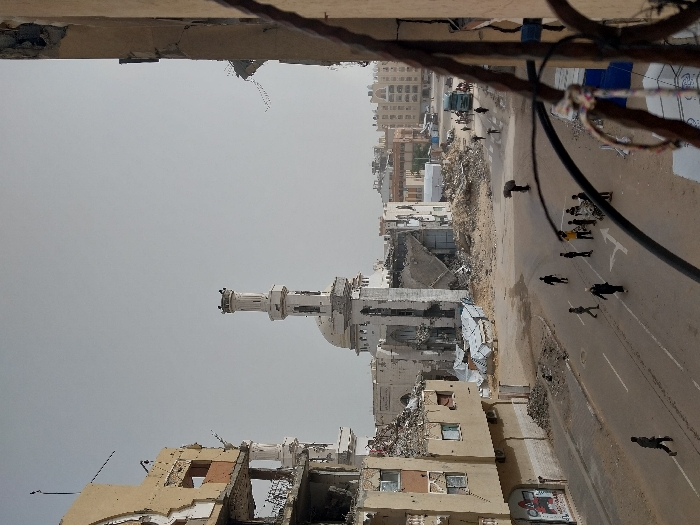 The image presents a severely damaged urban landscape in Khan Yunis, Palestinian Territory, under an overcast sky. In the central background, a mosque with a prominent, relatively intact minaret shows extensive structural collapse, with large sections of its dome and main edifice missing, exposing internal elements. Surrounding the mosque and extending throughout the scene are significant accumulations of rubble and debris.
To the left, a multi-story building exhibits catastrophic damage, including a blown-out facade revealing destroyed interior spaces and large piles of rubble on its remaining floors and at its base. Graffiti in red and black is visible on a lower section of this building. The right foreground shows the partial view of another damaged structure with exposed rebar.
A paved road with faded white lane markings (solid line, dashed line, and a directional arrow) cuts across the mid-ground. Approximately 15 individuals, appearing to be adults and children, are scattered across the street and the dusty, debris-laden adjacent areas. One person in the mid-ground is depicted carrying a large bundle on their back. A group of individuals is gathered in the center of the road. In the mid-distance to the right, a large truck is visible on the road. The ground surfaces are largely covered in dust, pulverized concrete, and miscellaneous building wreckage.