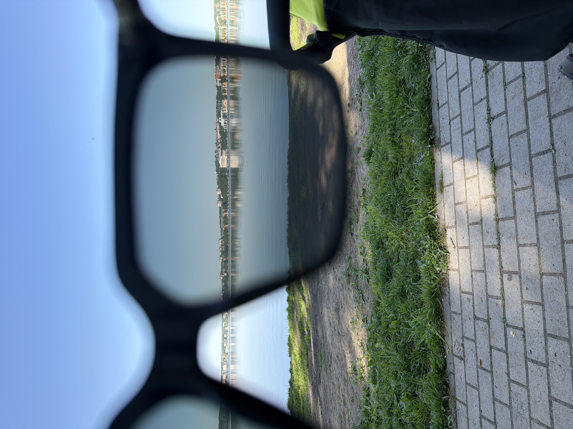A person stands on a brick path next to a patch of grass, holding up a pair of sunglasses that reflect a body of water and the opposite shoreline with buildings. The buildings on the shoreline suggest an urban or suburban setting. The image orientation is such that the ground and path are on the right side of the frame, while the sky reflected in the sunglasses occupies the left. The angle of the shot is slightly tilted. The scene is likely outdoors in daylight. The location is Votkinsk, Russia.