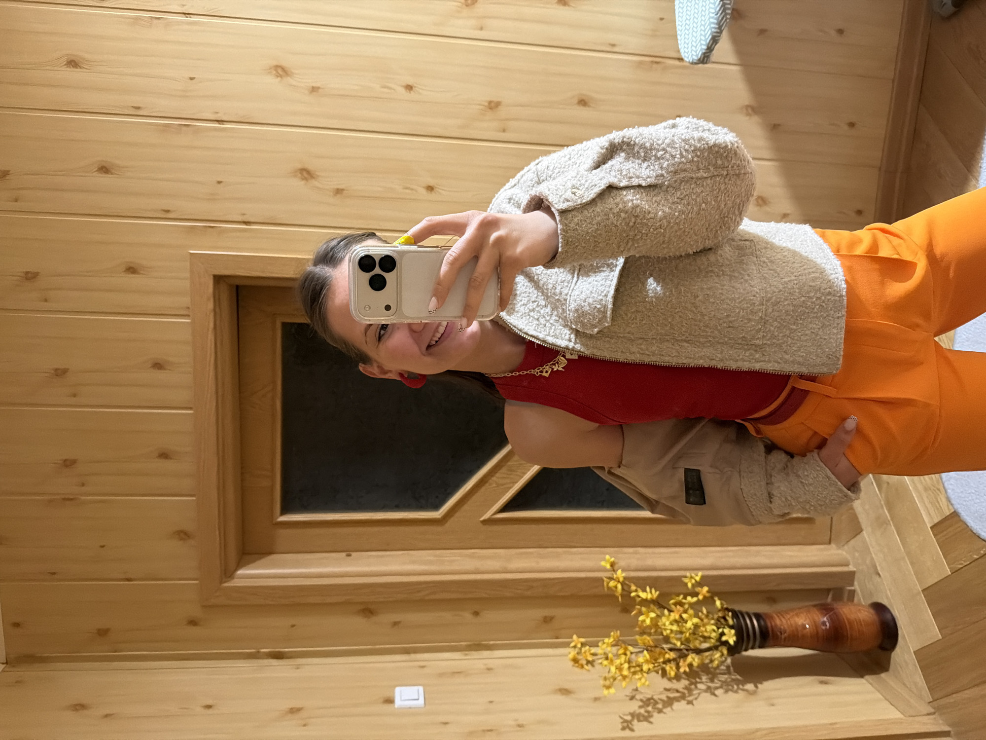 A young woman is taking a selfie indoors against a wall made of light-colored wood paneling, which suggests a cabin-like environment. She is holding a smartphone, with the camera facing her, and smiling. She is wearing a red top, an off-white fuzzy jacket, and orange pants. A gold necklace is visible around her neck. A framed opening, possibly a door, is behind her. A vase with yellow flowers is placed on the shelf beneath. The location cannot be verified as Buhovo, Bulgaria.