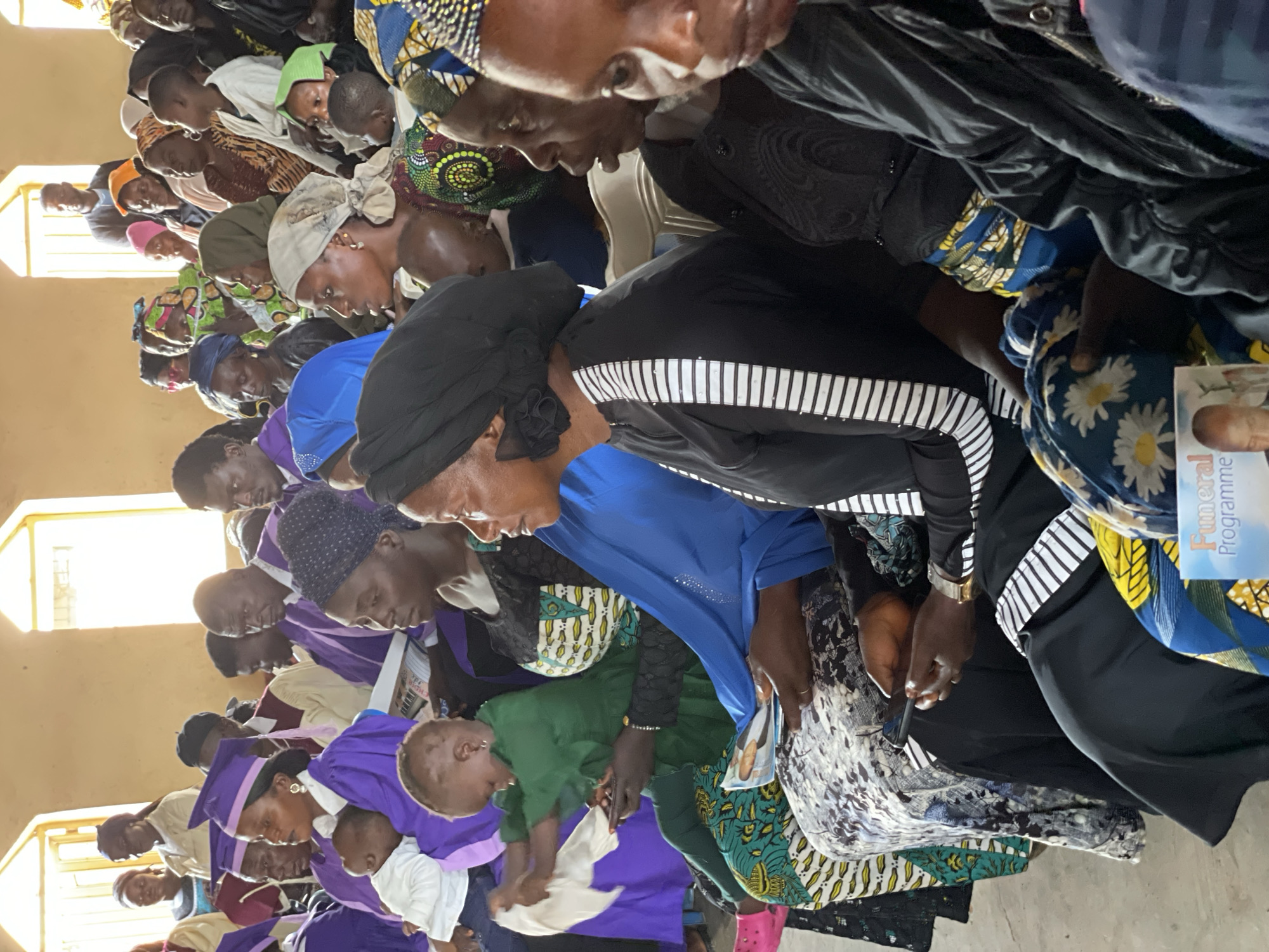 A group of people, likely attending a funeral service, are seated in an indoor venue in Kassa, Nigeria. The individuals are diverse in age and gender, with many wearing traditional Nigerian attire. Some are holding programs or documents, and some are using mobile phones. The room appears to be sparsely decorated, with natural light filtering in from windows. The overall atmosphere suggests a somber and reflective gathering.