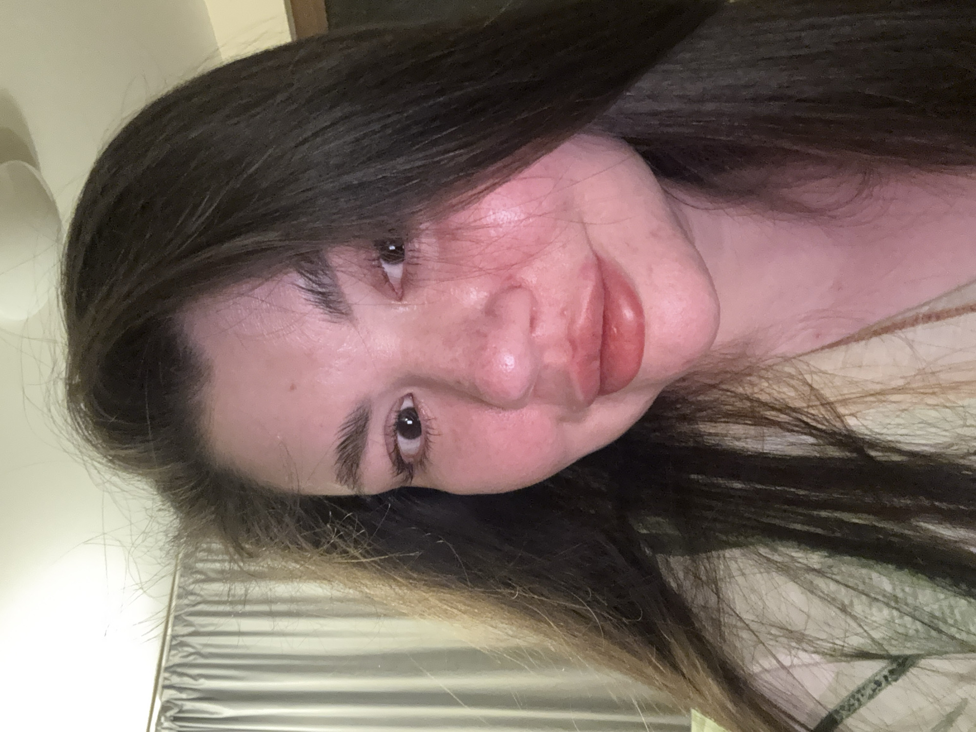 A close-up selfie of a woman with long, dark brown hair. Her skin appears fair, with a slightly flushed complexion on her cheeks. She has brown eyes and full lips. The background is indistinct, with blurred beige and gray tones. The location in the city of Votkinsk, Russia, cannot be determined from the image.