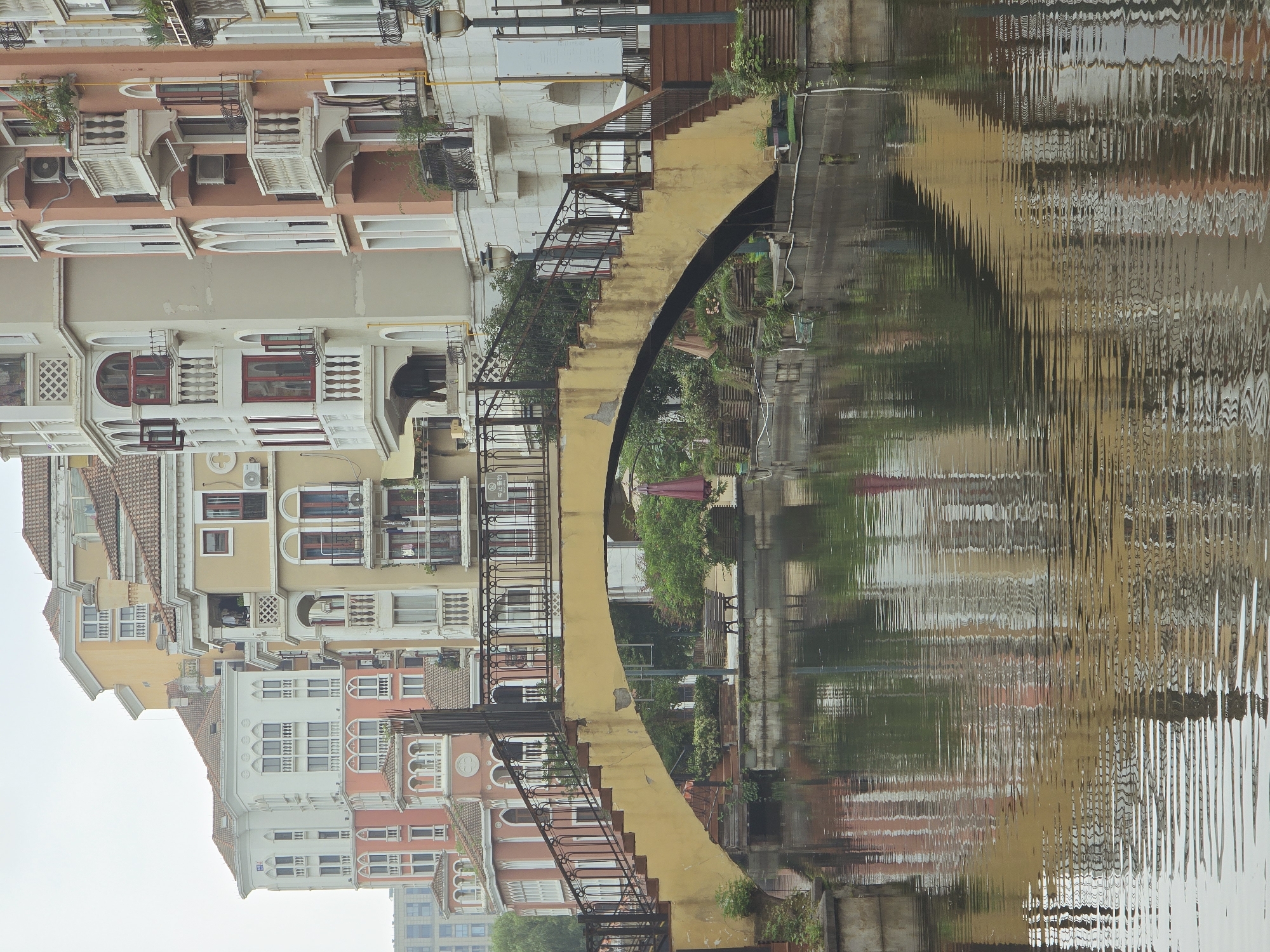 This is a medium-shot, eye-level, vertical photograph of a canal with a pedestrian bridge, located in the Xiaoshan District of China. The bridge, painted yellow and with a scalloped arch, spans the canal. Dark metal railings line the bridge's walkway, which is bordered by a retaining wall and a row of buildings. The buildings are multi-story structures with a European architectural style, featuring terracotta roofs, balconies with decorative railings, and arched windows in various colors including white, yellow, and pink. The canal's surface reflects the buildings and the bridge, creating a distorted image due to ripples on the water. Greenery, including trees and potted plants, is visible along the canal banks and on some of the building balconies. The sky is overcast, casting a diffused light on the scene. There are no people visible in the frame.