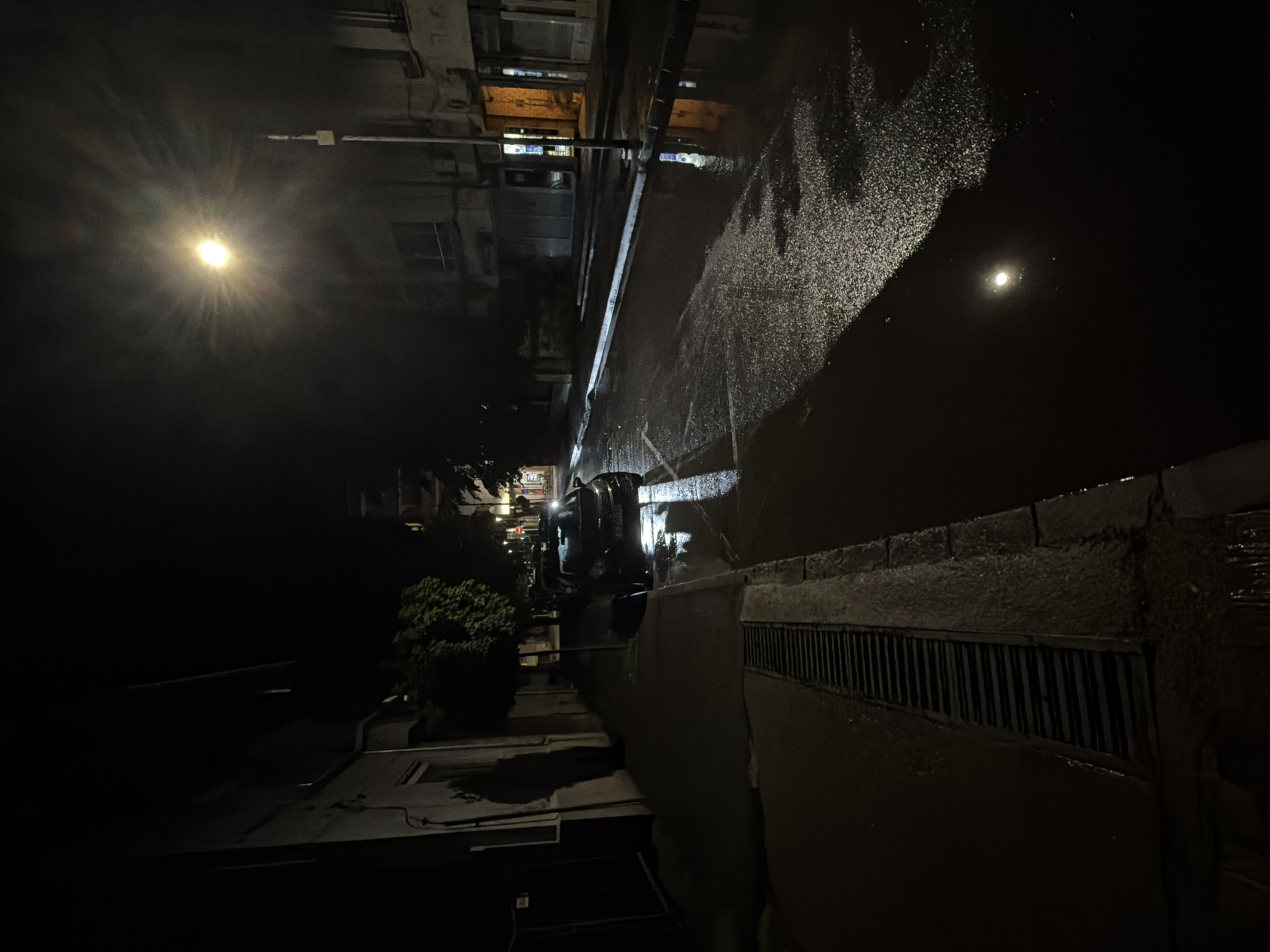 An outdoor, nighttime scene captures a wet street or alleyway in Pirgovo, Bulgaria, likely after rain. The image depicts a dimly lit urban environment under dark skies. On the left side of the frame, a bright street lamp emits a strong, star-like burst of light, serving as the primary light source for the foreground. The paved ground, possibly asphalt, is visibly wet and highly reflective, mirroring the street lamp and scattering speckled light from other sources across its surface.
Buildings line the right side of the street, with faint light emanating from some windows, suggesting interior illumination. Further down the street, a dark-colored vehicle, possibly an SUV or van, is parked or driving, partially obscured by the low light conditions. Green foliage from trees or bushes is visible on the left side, behind the street lamp, providing a touch of nature amidst the urban setting. A prominent metal storm drain or grate runs along the lower right edge of the frame, bordering the wet surface of the street or sidewalk.
No people are visible in the scene, and no specific activity is taking place, contributing to a quiet, almost still atmosphere characteristic of a post-rain night in a residential or urban area. There is no visible text in the image.