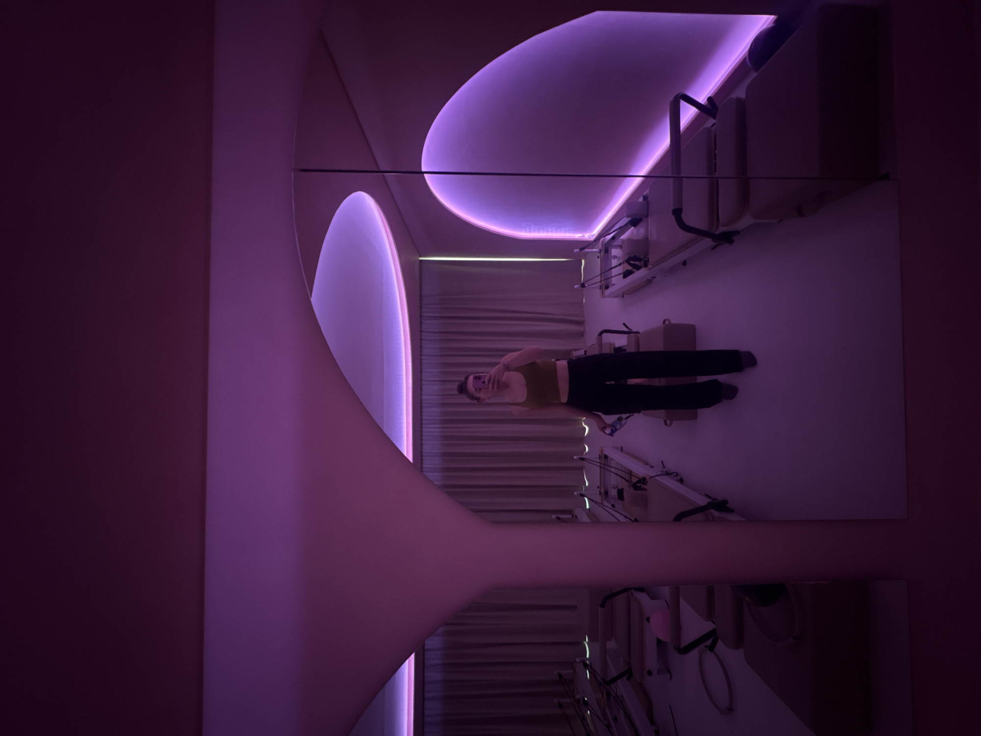 The image shows the reflection of a person in a Pilates studio, possibly in Mexico City, Mexico. The individual is standing on a Pilates reformer machine, wearing a tank top and dark pants. The room is lit with purple ambient lighting, creating a futuristic or modern aesthetic. The reflection shows the ceiling with curved architectural elements and additional Pilates equipment lining the walls. The studio appears to be well-equipped, suggesting a professional setting.