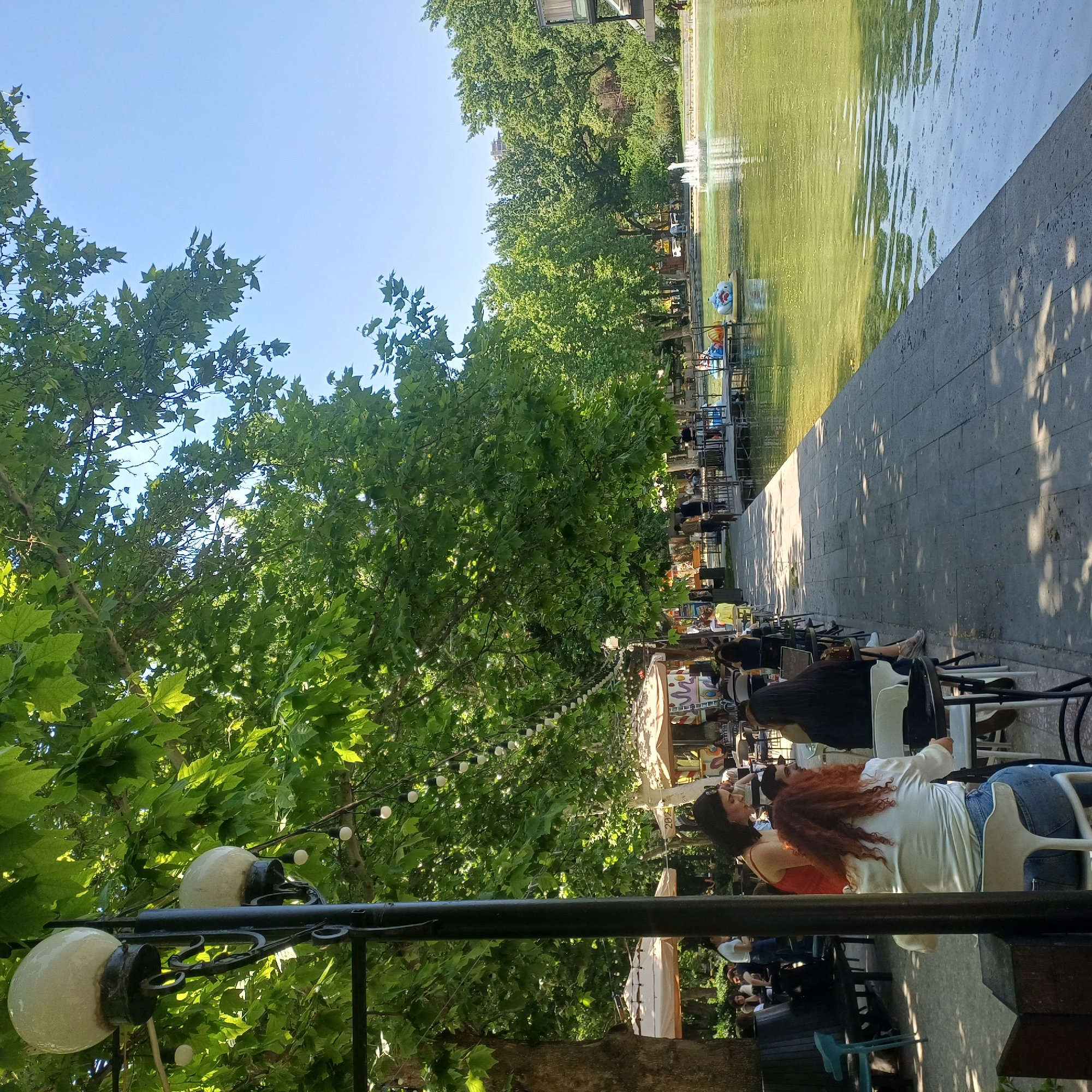 A street lamp with two globe lights stands in the foreground, to the left of the image, casting a shadow on a paved walkway. Beyond the walkway is a body of water with a fountain in the middle and several pedal boats docked on the edge. Trees with lush green foliage line the walkway and the opposite bank of the water, casting dappled shadows. Several people are seated at tables under umbrellas on the left side of the walkway, appearing to be in an outdoor cafe. The sky is clear and blue.