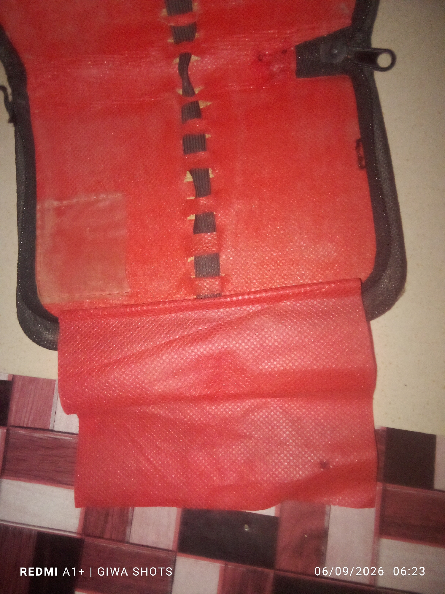 A close-up, overhead shot of an open red fabric case with black trim is pictured. The case appears to be a storage pouch for something, possibly a cosmetic bag or a small tech accessory holder. The interior of the case is lined with a red fabric that has a fine, dot-like texture. A section of the case is folded over, revealing a second layer of red fabric with a similar textured pattern. The edge of the case features a black zipper with a circular pull tab. The case is placed on a surface with a tiled pattern, composed of squares in shades of brown, white, and black. The bottom left corner of the image shows white text that reads "REDMI A1+ | GIWA SHOTS," and the bottom right corner displays a timestamp: "06/09/2026 06:23." The overall lighting is dim, and the image quality is slightly grainy.