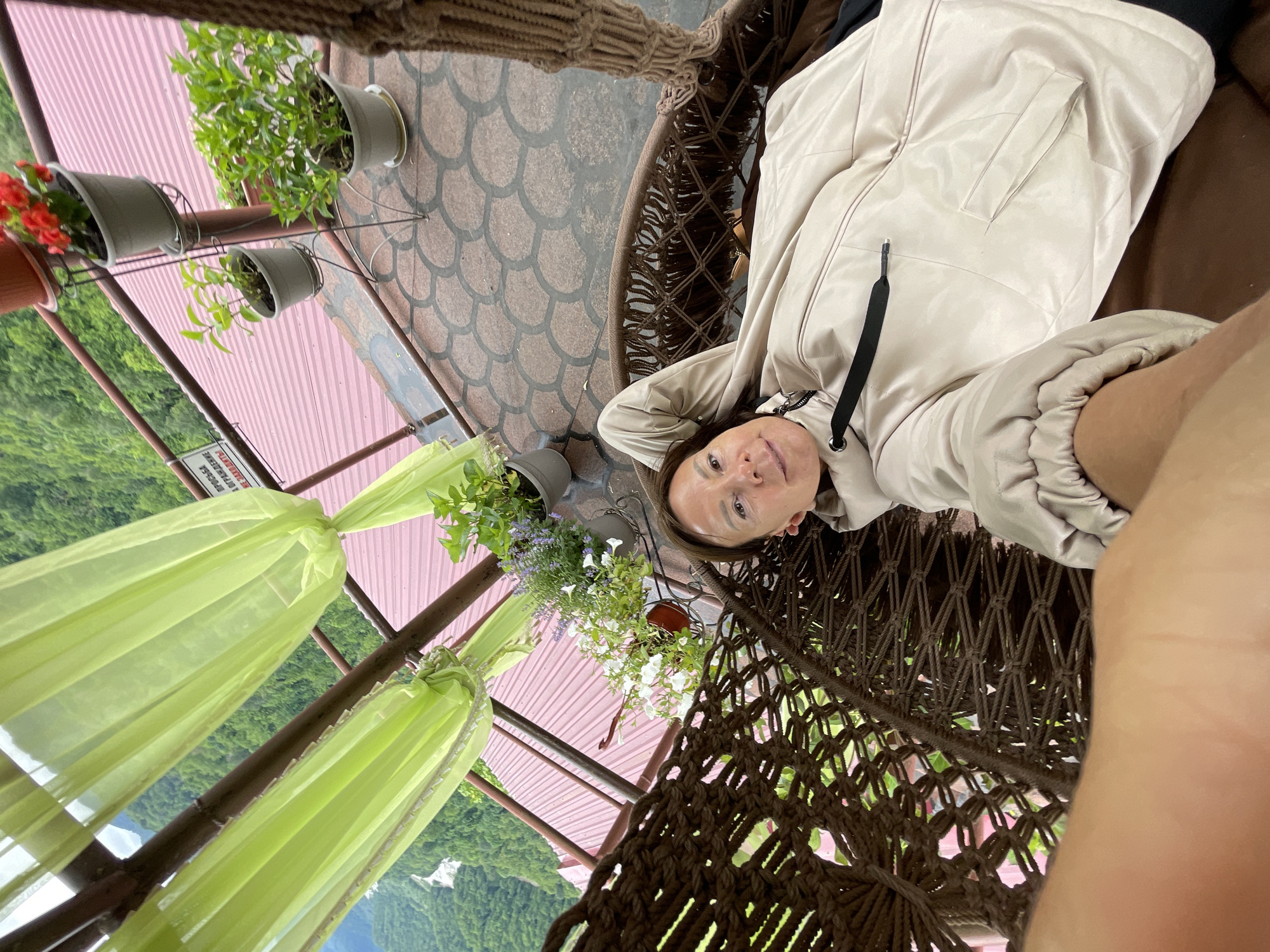 A woman in a light-colored jacket is in a hanging chair, possibly on a porch with greenery and flowers. The location is not visible in the image.