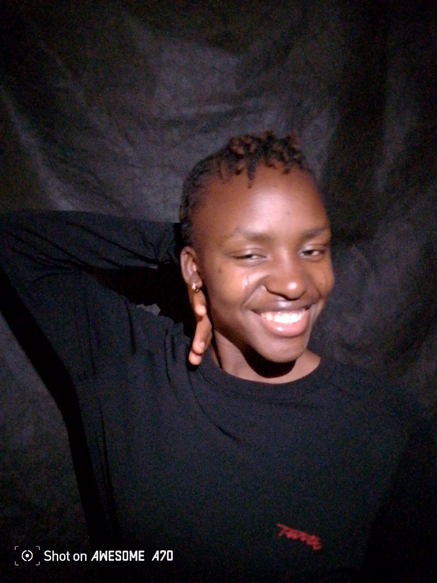 A medium shot shows a young woman with dark skin smiling and looking directly at the camera. She is wearing a dark long-sleeved shirt. Her left arm is raised behind her head. She appears to have short, braided hair. The background is dark and appears to be a textured black cloth or backdrop. The bottom of the frame has text that reads "Shot on AWESOME A70". The location is not identifiable.