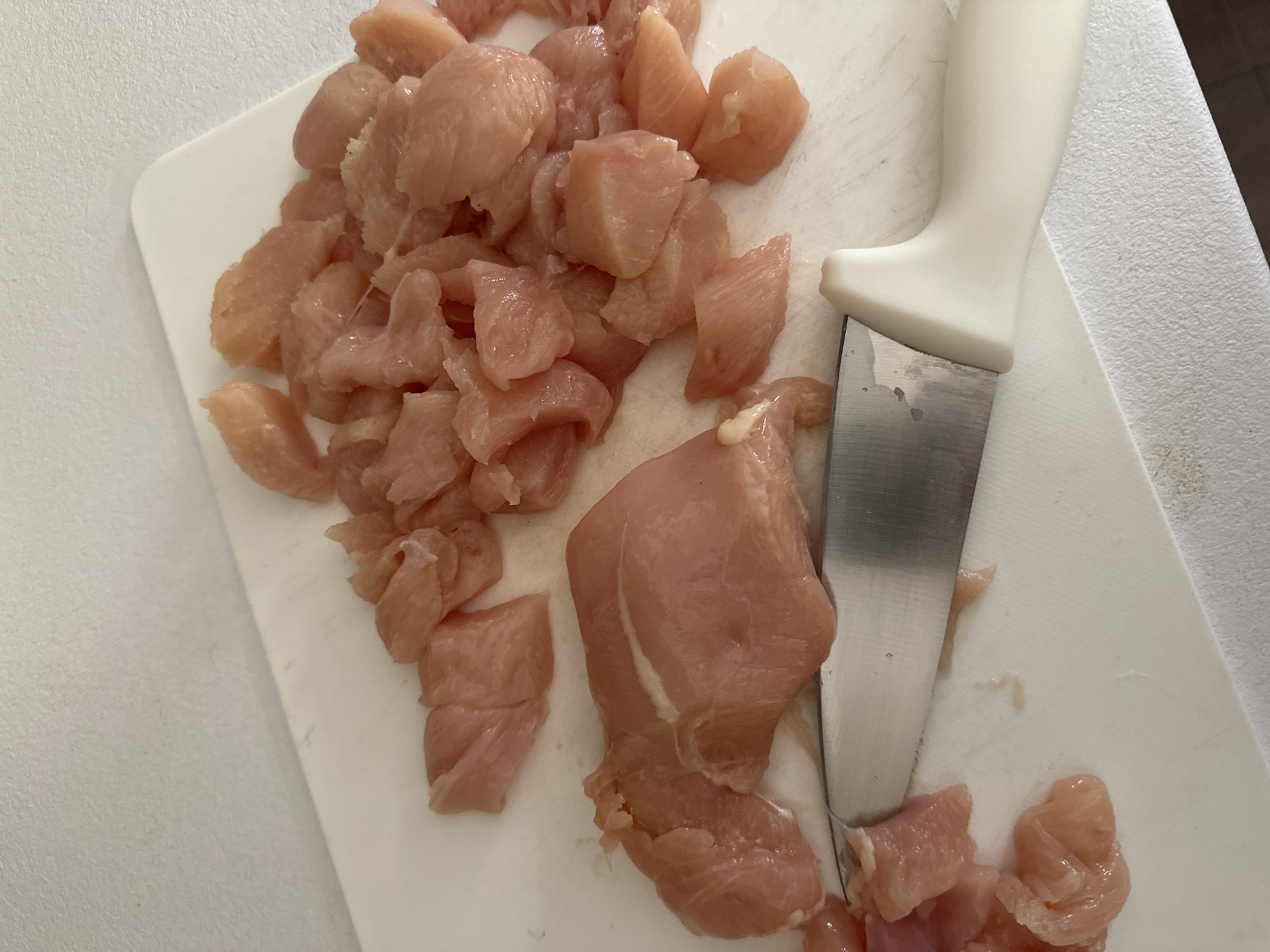 The image shows diced raw chicken on a white cutting board, partially cut with a knife that has a white handle. The scene appears to be indoors, possibly in a kitchen. The location within Budva, Montenegro cannot be determined from the image.