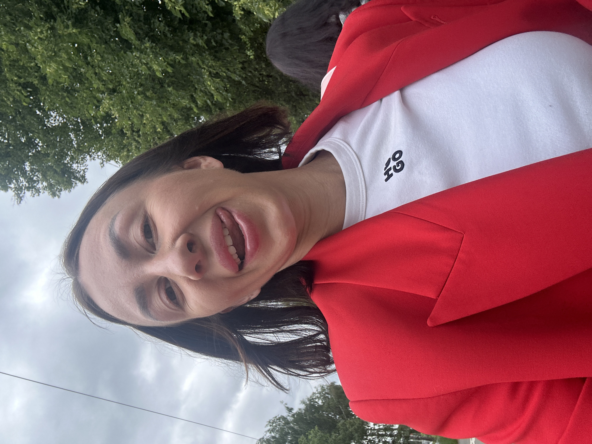 A woman in a red blazer and white t-shirt with a "HUGO GO" logo is taking a selfie outdoors. She is sticking her tongue out and smiling. Her hair is dark and shoulder-length. In the background, there are trees and a cloudy sky. A portion of another person's dark hair is visible to the right. The scene appears to be on a street or sidewalk in a city, likely Votkinsk, Russia, given the context.