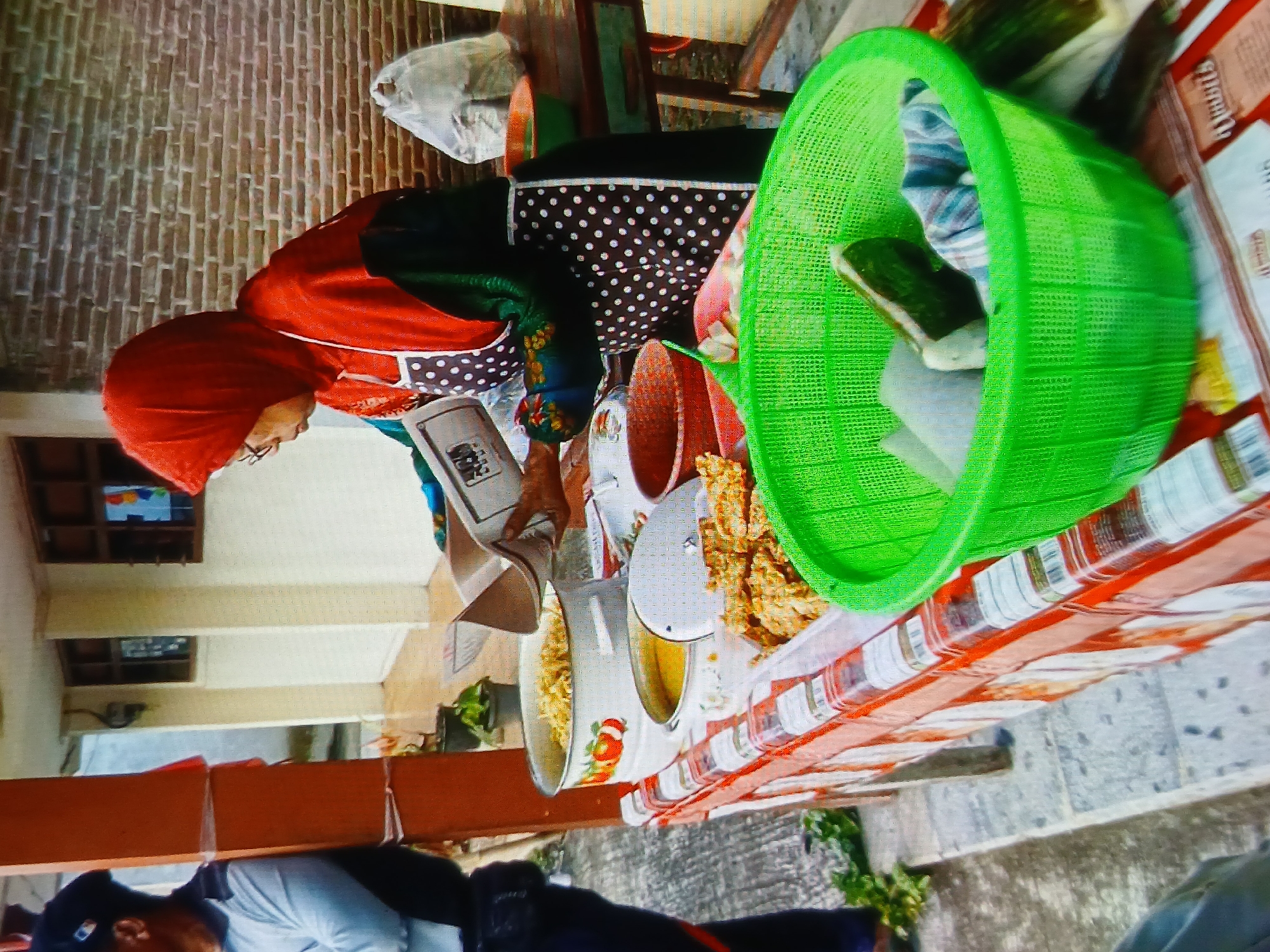 A street vendor wearing a red hijab and a black and white polka dot apron is weighing food items on a scale. She is standing behind a table laden with various food dishes, including what appears to be fried tempeh and noodles in pots. A bright green plastic basket filled with food items sits in the foreground. Another person, partially visible on the left, is also present. The scene is set outdoors, with a brick wall in the background and what appears to be a tiled floor or walkway. The overall setting suggests a food stall or market in Setia Bakti, Indonesia.
