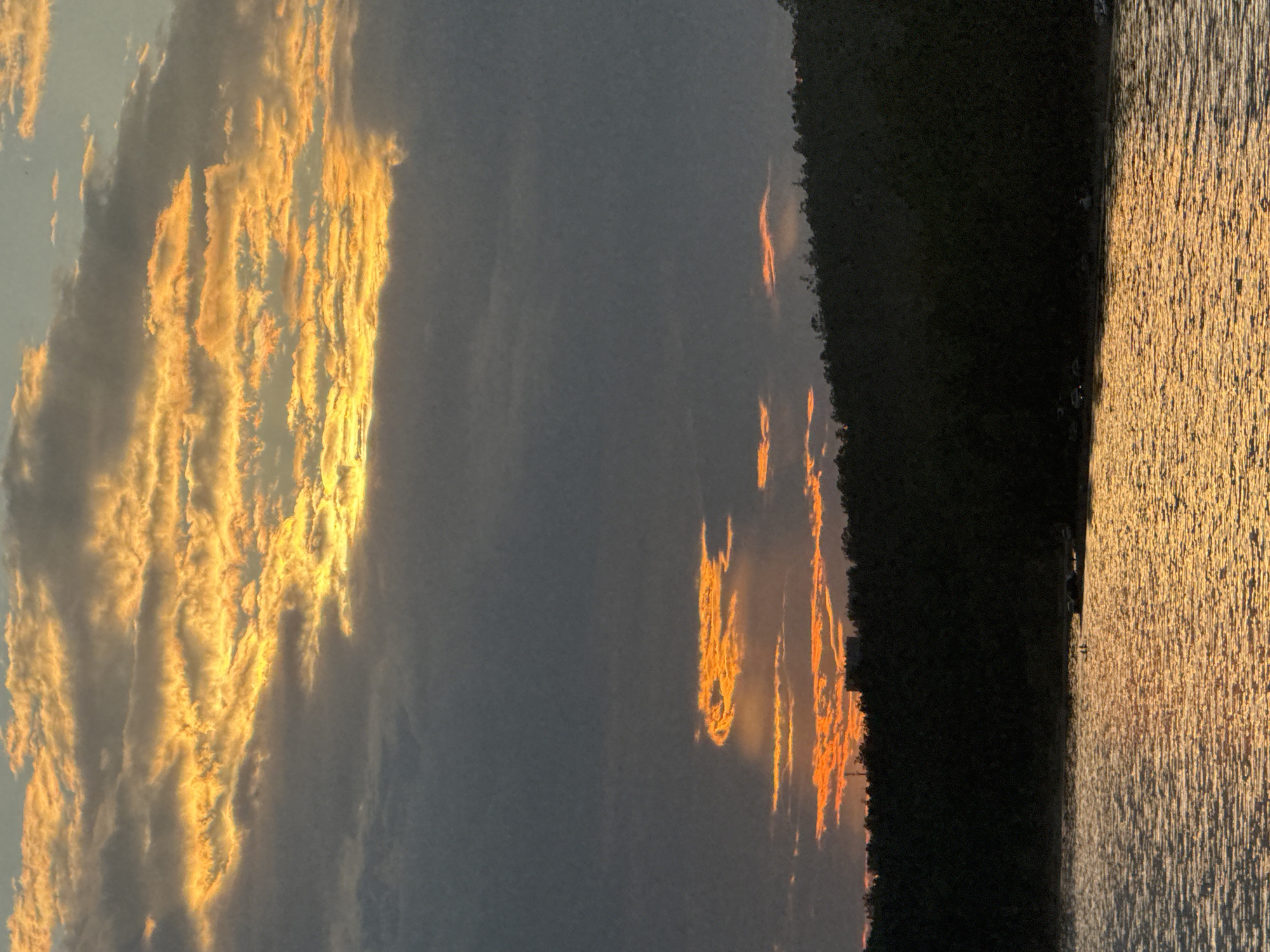 This is a photograph of a body of water reflecting the colors of a sunset. The water is in the foreground, with a dark, tree-covered hill forming the horizon. Above the hill, the sky is filled with clouds, some of which are illuminated by the setting sun in hues of orange and yellow. The overall impression is one of a tranquil natural scene. The image is likely taken in Bratsk, Russia, given the context. There are no visible people, animals, or specific man-made objects that would indicate the exact location within the city, but the presence of water and trees suggests a natural setting that could be found within or near Bratsk. The time of day is sunset.