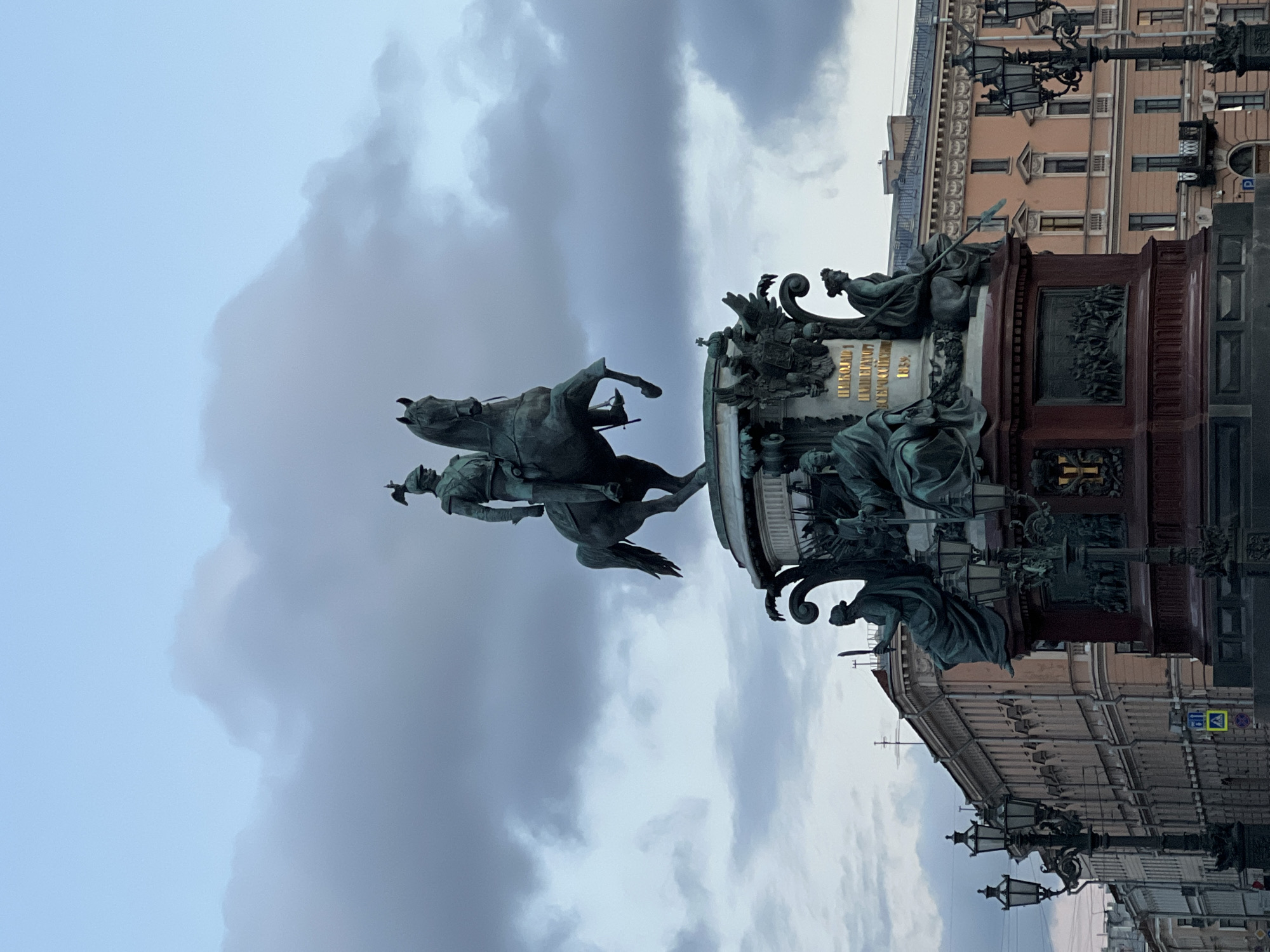 The image shows the equestrian statue of Nicholas I in Saint Petersburg, Russia. The monument stands prominently against a cloudy sky. The statue depicts Nicholas I on horseback, with figures surrounding the base of the monument. Buildings and ornate street lamps can be seen in the background.