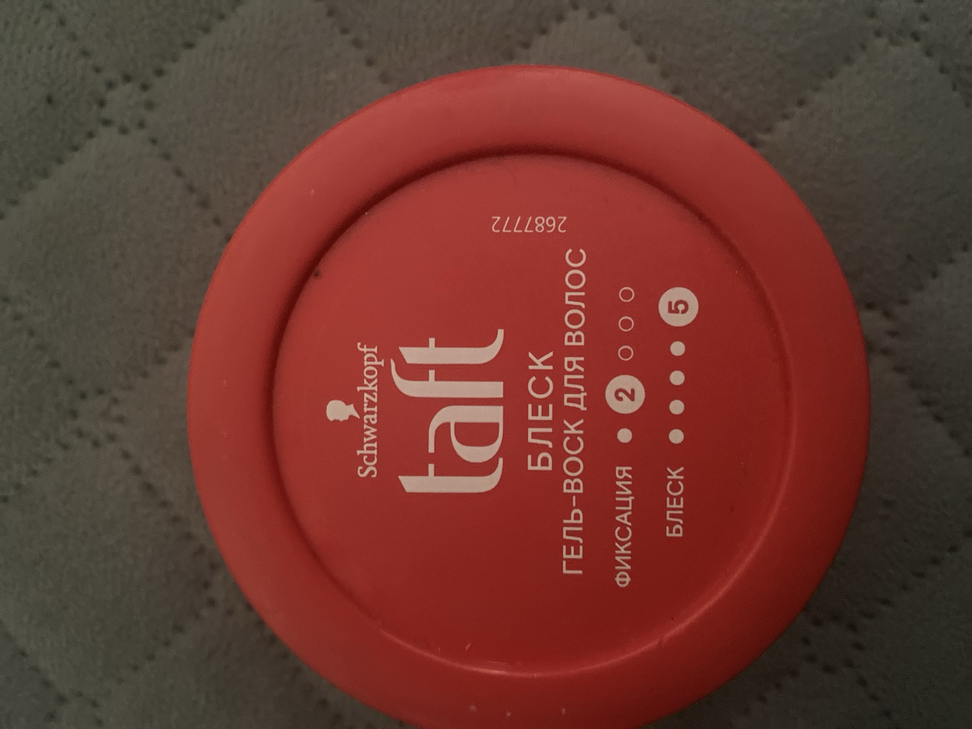 A red, circular plastic container with white text is pictured from a top-down perspective. The container is placed on a dark, textured surface, possibly a bedspread or carpet. The lid of the container features the brand name "Schwarzkopf" above the logo "taft" in large white letters. Below this, in smaller white text, are the words "БЛЕСК" (Gloss) and "ГЕЛЬ-ВОСК ДЛЯ ВОЛОС" (Gel-Wax for Hair). Further down, there are indicators for "ФИКСАЦИЯ" (Fixation) with a dot next to it and a circled number "2," and "БЛЕСК" (Gloss) with three dots and a circled number "5." A serial number "2687772" is printed vertically on the right side of the container lid. The image does not contain any identifiable elements related to the city of Kizilyurt, Russia, or its specific location. There are no people or actions depicted in the image.