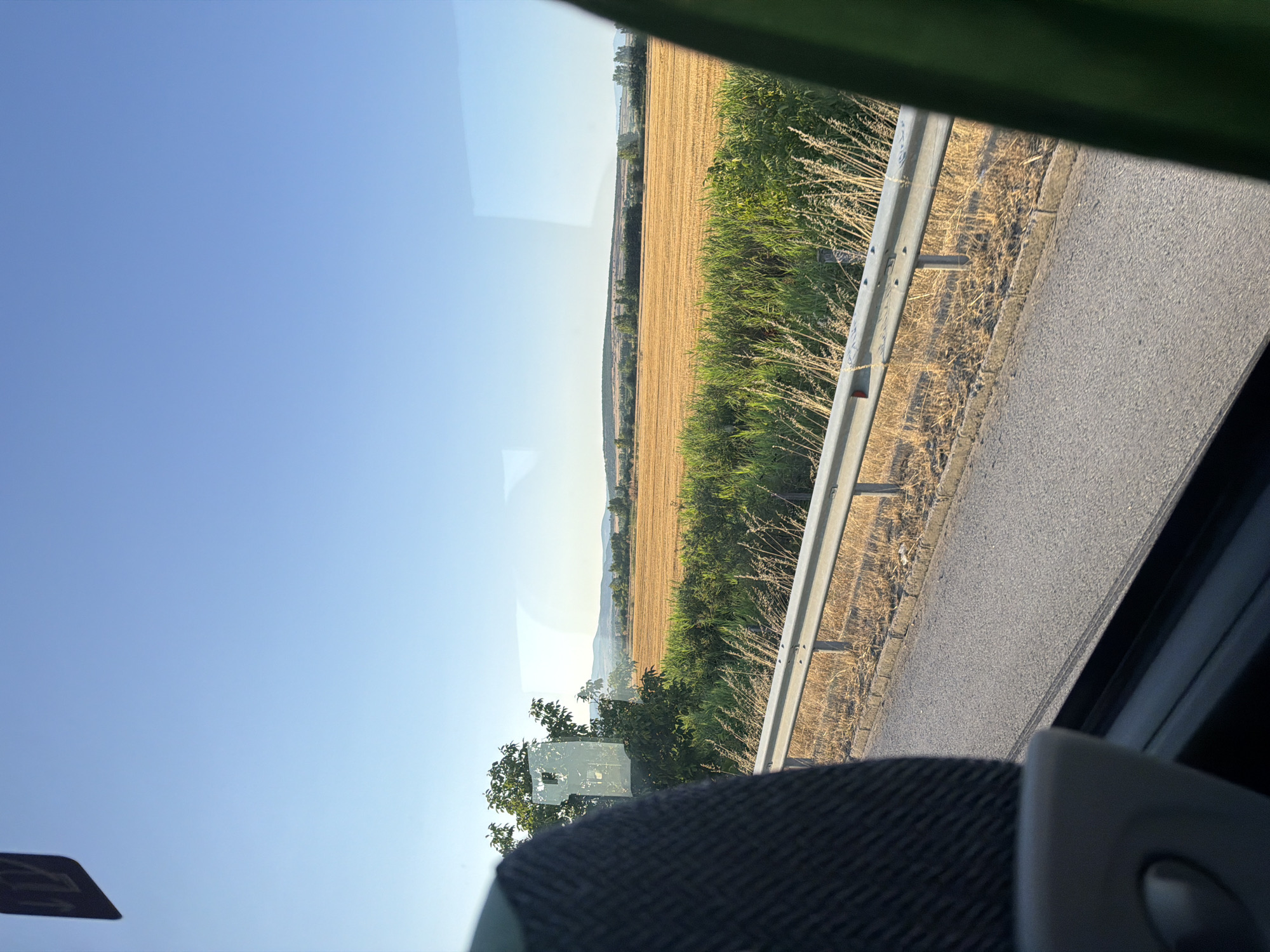 The image shows a view from inside a vehicle, likely a bus, looking out a window. In the foreground, the dark, textured fabric of a seat is visible on the left, and a portion of a green material is on the right. A metal guardrail runs along the side of a paved road, which is partially visible in the lower portion of the image. Beyond the road and guardrail is a dry, golden field, likely a harvested crop, stretching into the distance. Green vegetation, including tall grass and some bushes, borders the field. In the background, a line of trees and rolling hills can be seen under a clear, bright blue sky. The scene appears to be set in a rural area, possibly the vicinity of Shtarkovo, Bulgaria, given the landscape and the time of day suggesting a sunny afternoon. There are no people or animals clearly visible in the image.