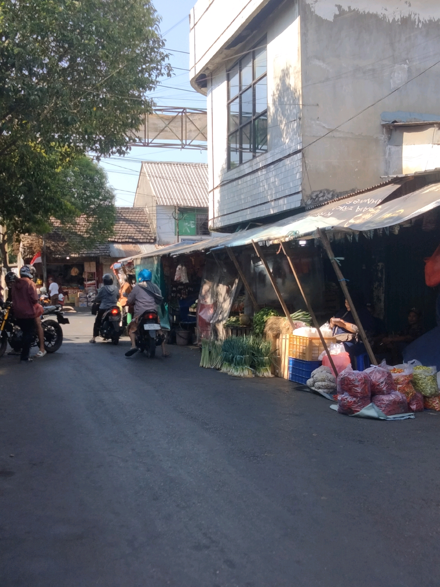 This is a vertical, eye-level shot of a street scene in Mojokerto, Indonesia, likely a market area. The foreground is dominated by a paved street. To the right, a market stall is set up under a tarp, displaying various produce including large bunches of green onions, bags of red chili peppers, and other vegetables. Two women appear to be seated within the stall, one of whom is looking towards the camera. In the background, several people are riding motorcycles on the street, and further back, buildings with tiled roofs are visible. A large green tree is present on the left side of the image, with overhead power lines or infrastructure crossing the frame. The lighting suggests it is daytime, with sunlight casting shadows.