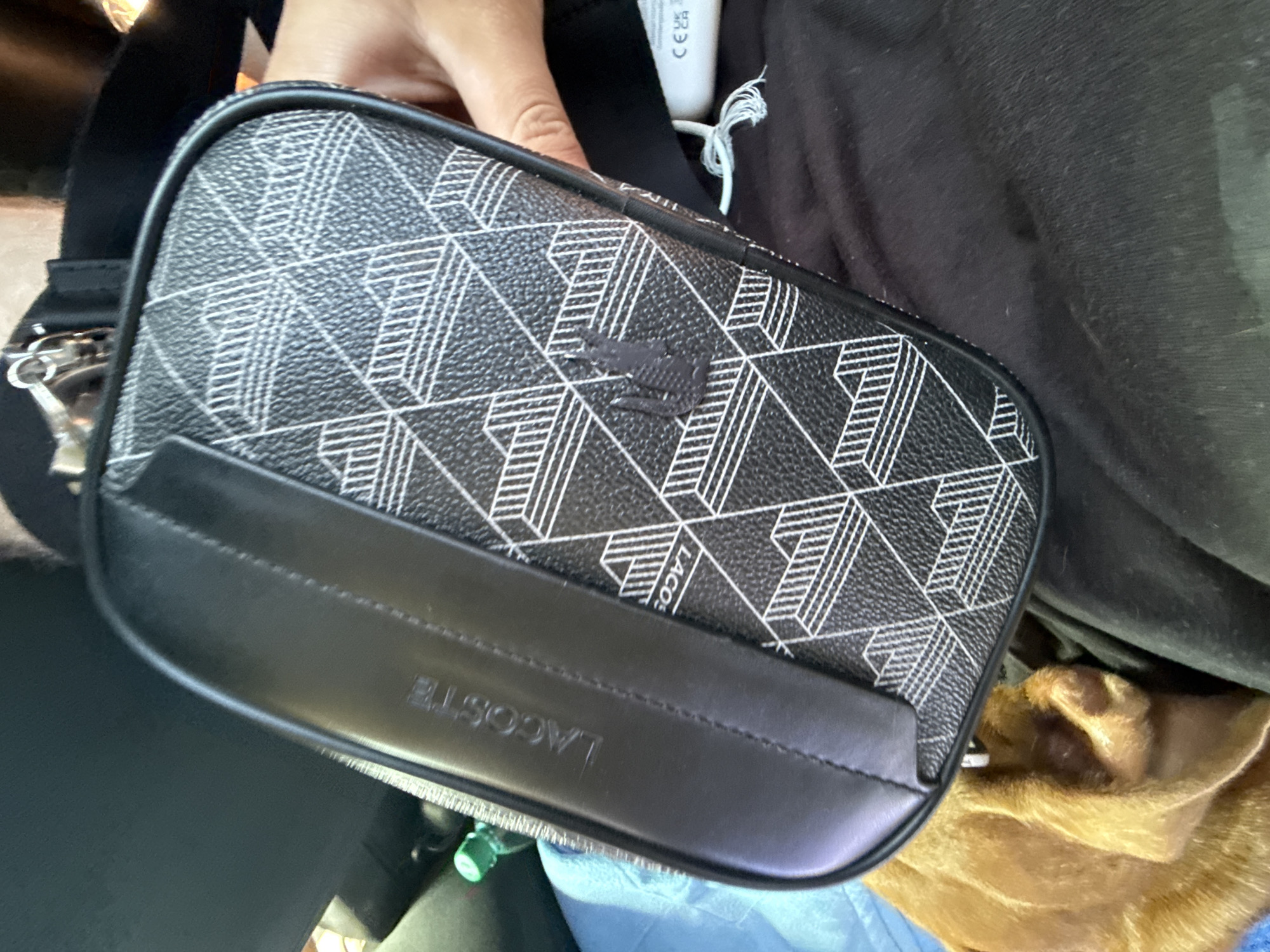 The image shows a close-up view of a black and white geometric pattern toiletry bag with the "LACOSTE" logo embossed on a black leather strip. The bag has a small, dark embossed crocodile logo on its patterned surface. A person's hand is visible on the right side of the image, holding the bag. The bag is placed on a dark fabric surface, possibly clothing, and a small part of a brown dog is visible at the bottom left. In the background, a partially visible green bottle is present. There is no indication of the city of Vyzyrka, Ukraine, in the image.