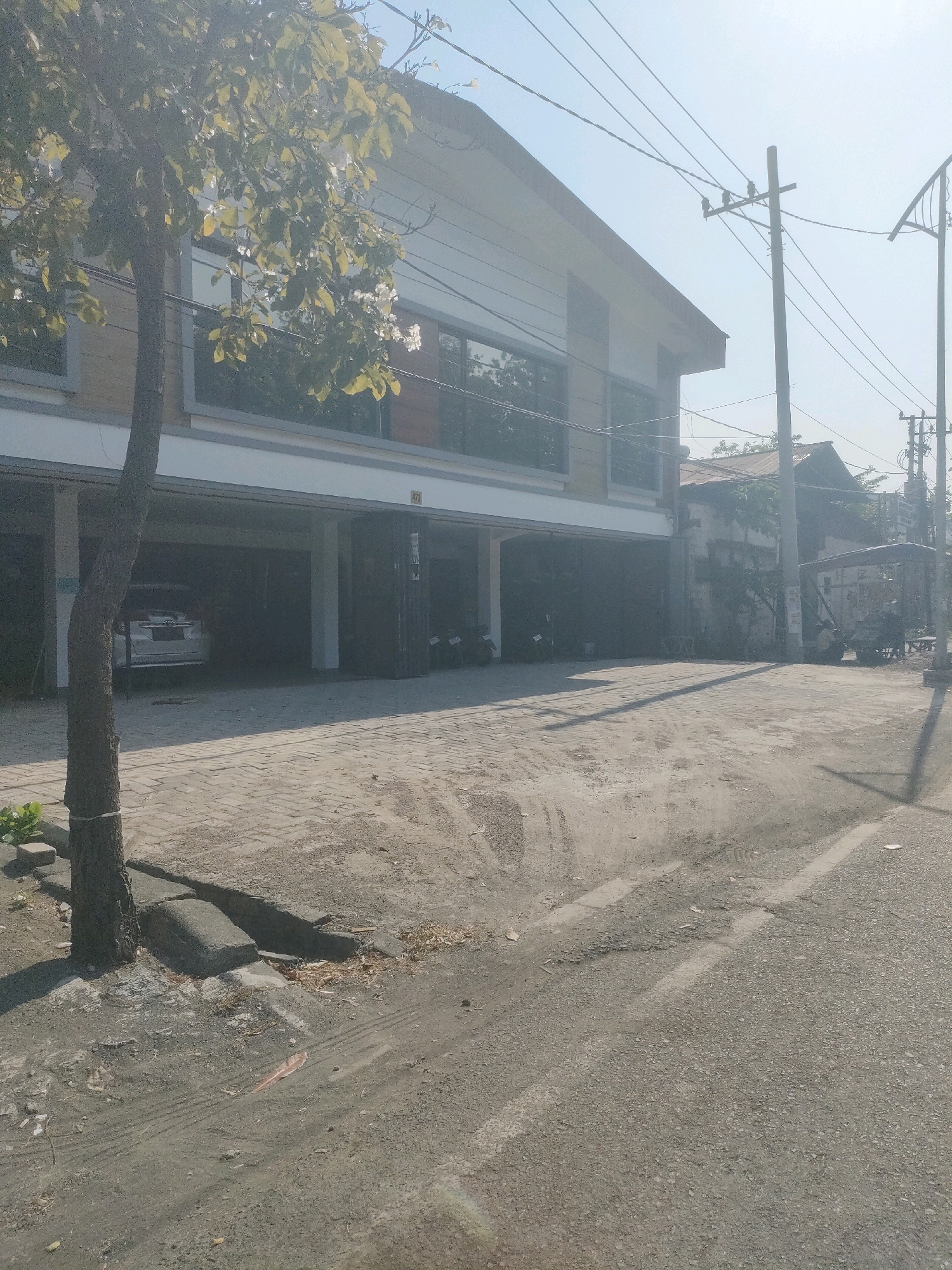 The image depicts a street scene in Surabaya, Indonesia, on a bright, sunny day. In the foreground, a paved road transitions to a brick-patterned sidewalk with a tree on the left. The tree has a rope tied around its trunk. A small green plant is visible near the base of the tree. A concrete curb runs along the edge of the sidewalk.
Behind the sidewalk is a two-story building. The ground floor of the building appears to be open, resembling a parking or commercial space, with a car parked inside on the left. The upper floor has large windows with dark frames, reflecting the sky and trees. Electrical wires and utility poles are prominent in the background, stretching across the sky.
Further in the background, other buildings with tiled roofs are visible, along with more trees. A cart-like vehicle is parked on the right side of the street. The overall environment suggests an urban or semi-urban setting, possibly a commercial area or a residential street with businesses. The lighting indicates it is daytime, with shadows cast by the buildings and the tree.