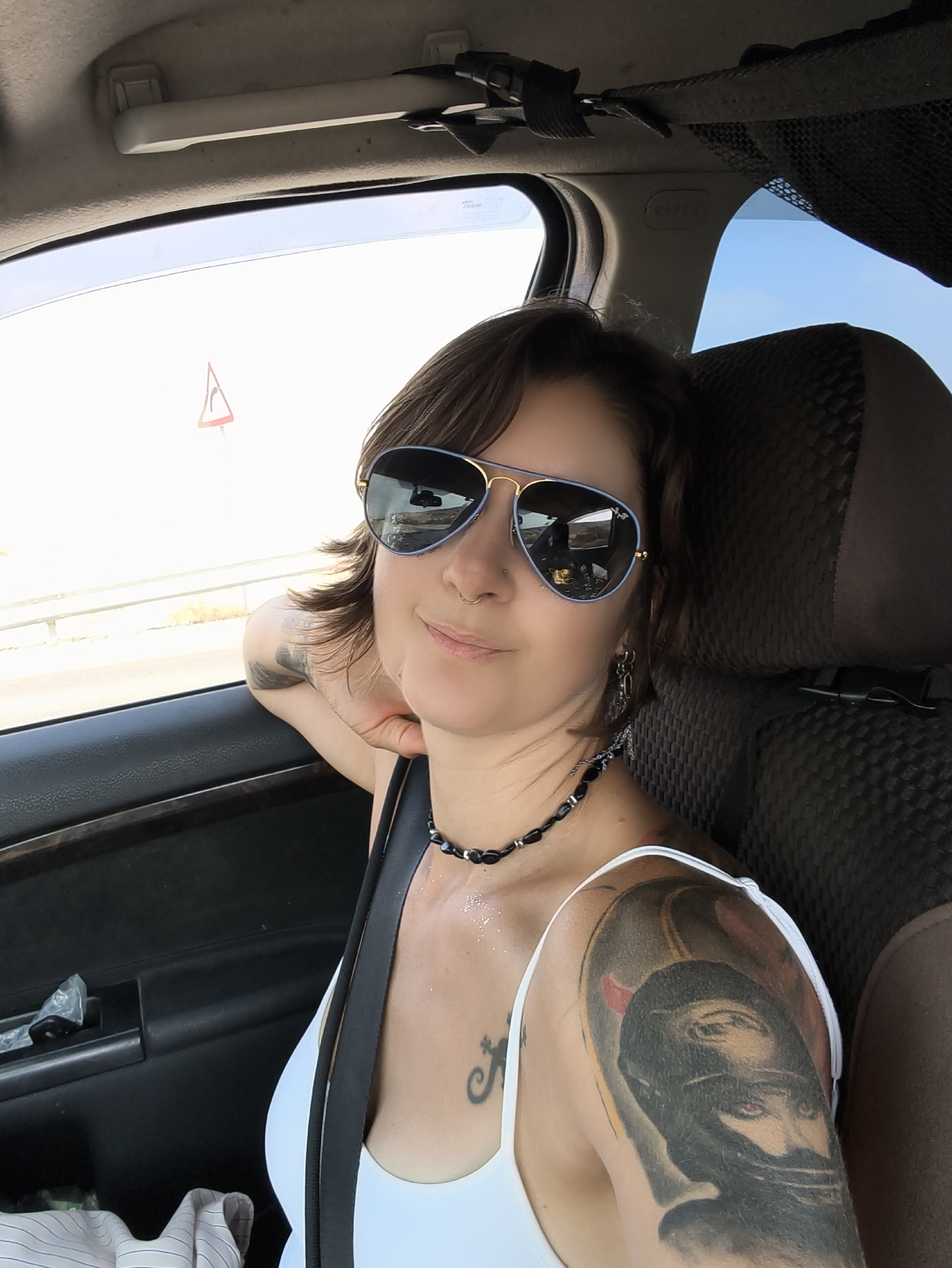 A woman is seen inside a car, looking towards the camera and smiling. She is wearing aviator sunglasses, a black beaded necklace, and a white tank top. She has tattoos on her left arm and shoulder. The woman is in the passenger seat of the car, which appears to be driving on a road, indicated by the road sign visible through the window. The scene is likely in Ramat Negev Regional Council, Israel, given the context. The interior of the car shows the seat and door panel. The lighting suggests it is daytime.