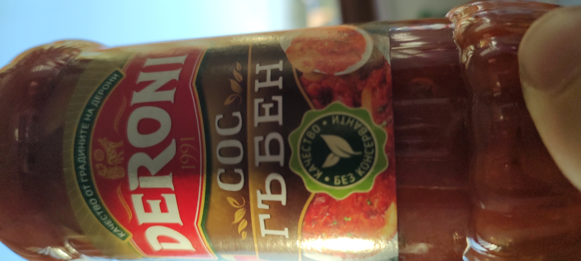 The image shows a jar of "Deroni" brand tomato sauce with cheese. The label includes the year "1991" and the text "СОС ГЪБЕН" which translates to "Cheese Sauce". A green seal on the label states "КАЧЕСТВО ГАРАНТИРАНО БЕЗ КОНСЕРВАНТИ", translating to "Quality guaranteed without preservatives." The jar is in focus, with parts of the background and a finger slightly blurred. The location is not identifiable from the provided image.