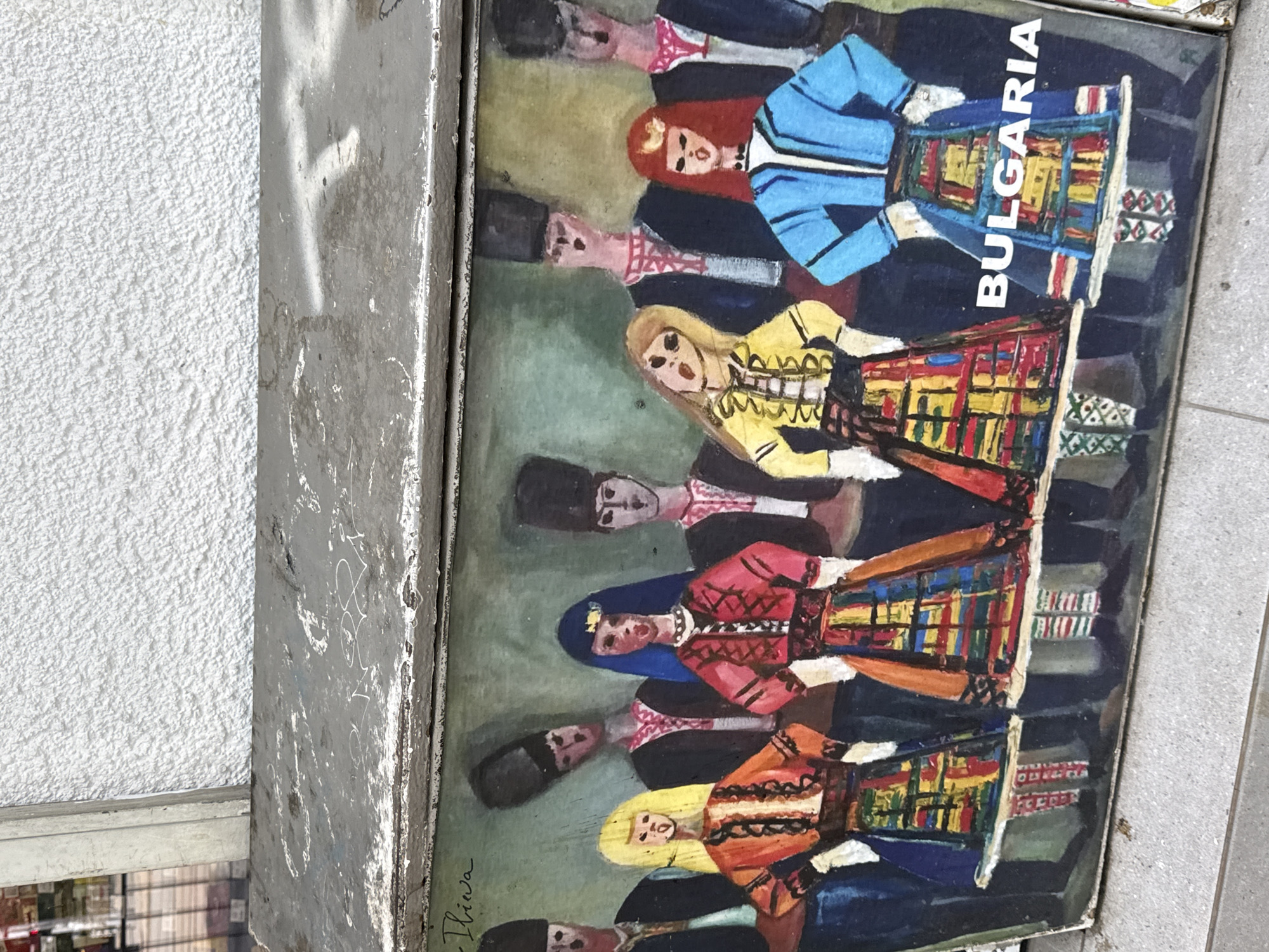 The image displays a vertically oriented painting, seemingly a print or reproduction, depicting several figures in traditional Bulgarian folk costumes. The painting is housed in a weathered grey metal frame and is shown in an outdoor setting.
Within the painting, male and female figures are arranged in rows, all facing forward. The men wear dark hats and light-colored shirts with patterned details, while the women are dressed in vibrant, multi-colored embroidered dresses or skirts, vests, and often head coverings. The costumes feature intricate patterns in red, yellow, blue, and green. The figures are rendered in a folk art or naive style, with simplified features and bold colors.
Prominently featured in white capital letters, running vertically on the right side of the painting, is the word "BULGARIA." In the bottom left corner of the painting, there is a visible signature that appears to read "Iliwa."
The metal frame surrounding the painting shows significant wear, with chips, scratches, and some faded white marks or graffiti. To the left of the frame, there is a textured, off-white stucco wall, indicating an exterior building surface. To the right, a grey concrete or paved ground surface is visible. The lighting suggests it is daytime, with no strong direct sunlight.
The overall scene captures a cultural artifact, likely a souvenir or decorative piece, displayed in a public space in Sofia, Bulgaria, reflecting local heritage.