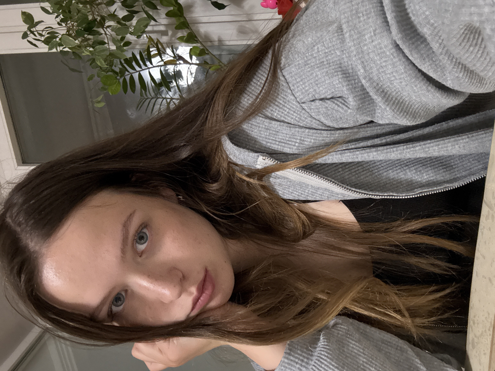 A young woman is seen in a close-up shot. She has long brown hair, light skin, and blue eyes. She's wearing a gray hooded sweatshirt. Behind her is a window with a plant partially obscuring the view. The location is Chaikovsky, Russia.
