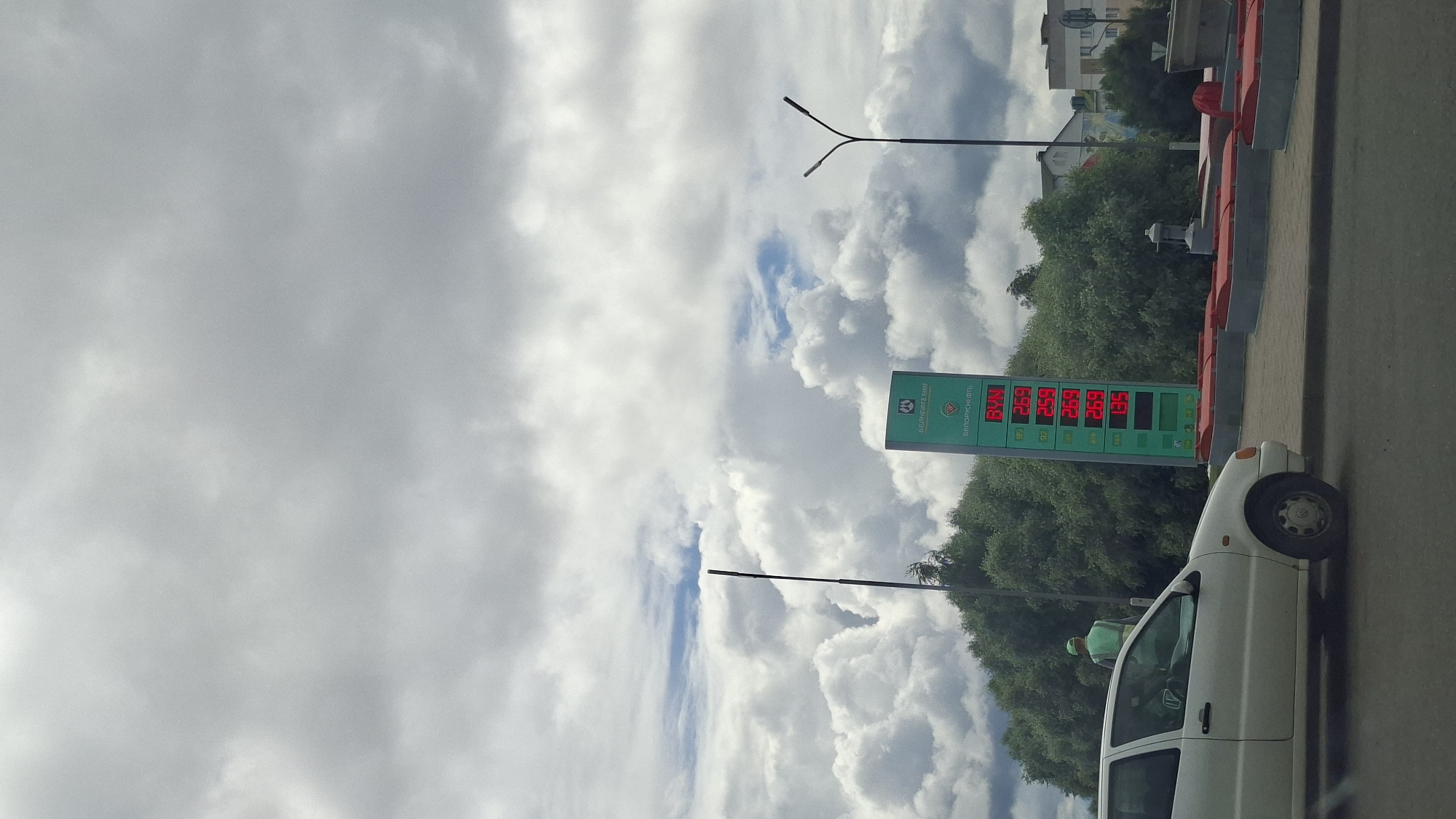 This is a vertical, eye-level, outdoor, full shot of a gas station price board and a white hatchback car. The gas station price board is green with red digital displays showing fuel prices. The prices displayed are 2.69 for 95 octane, 2.59 for 92 octane, 2.69 for 95 octane, 2.69 for AI-92, and 1.35 for diesel. Above the prices, the word "BYN" is displayed in red. The gas station's logo, which appears to be a green circle with white elements and text, is at the top of the board. The background consists of a large number of white, puffy clouds against a light gray sky. Below the clouds, there are lush green trees, and behind them, several buildings. A white car is parked in front of the gas station. A person wearing a light green vest and hat is visible inside the car. A dark pole is also present in the scene, standing tall against the sky and trees. The car is parked on a paved surface, likely a road or parking lot, with a red and gray curb in the foreground. The overall scene suggests a cloudy day in an urban or suburban environment in Myadzyel, Belarus.