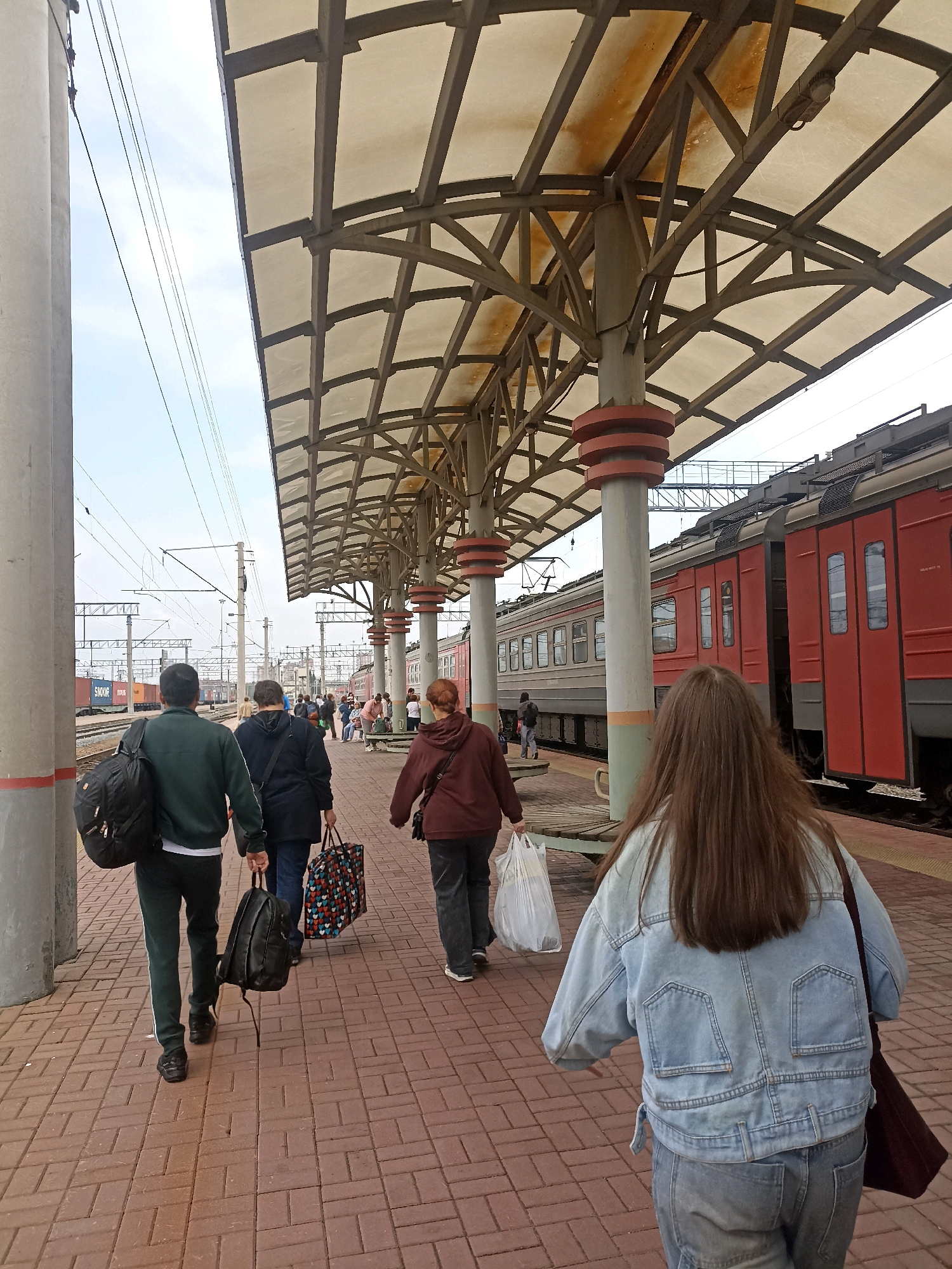 The image depicts a train station platform in Novosibirsk, Russia. Several people are walking along the platform, some carrying bags. A train is visible on the tracks next to the platform. The platform is covered by a canopy supported by pillars. The sky is overcast. The ground is paved with red brick. In the foreground, a person with long brown hair wearing a denim jacket and jeans walks away from the camera. To the left, two men in tracksuits are walking, one carrying a black backpack and the other a black duffel bag. Further down the platform, a woman in a maroon hoodie is walking with a plastic bag.