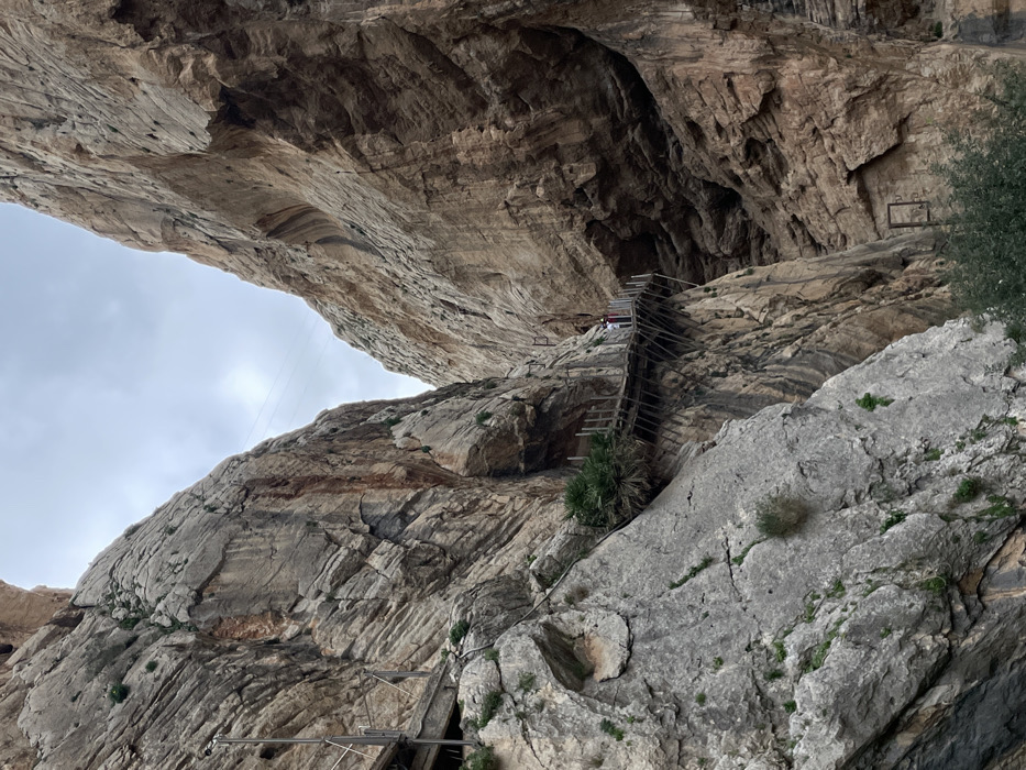 This image captures a dramatic view within a mountainous or canyon landscape, specifically featuring the renowned Caminito del Rey in El Chorro, Spain. The scene is dominated by massive, layered rock formations, showcasing steep cliffs and a pronounced overhang that creates a natural cavernous space overhead.
A prominent feature is the suspended wooden walkway or stairs, equipped with metal railings, that hugs the side of the cliff face, appearing to traverse under the large rock overhang. Several individuals are visible on this walkway; some are walking, while others appear to be seated or resting. Their presence highlights the scale of the rock formations and the precarious nature of the path.
The sky, visible through the opening between the rock formations, is overcast and bright, suggesting a cloudy daytime. There are no harsh shadows, contributing to the diffused light. Sparse vegetation, including small bushes and plants, clings to the rock surfaces, adding a touch of green to the otherwise rugged, earthy tones of the rock. In one of the cropped sections, a rectangular, empty frame-like structure is affixed to the rock face, possibly an old sign holder or marker. Cables, possibly for utilities or safety, are also faintly visible stretching across the void. No discernible text is present in the image.
