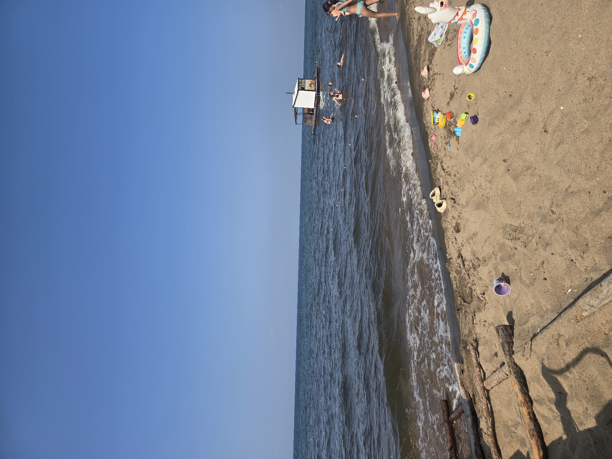 The image depicts a sunny day at a beach, likely in Maksimikha, Russia, given the provided context. The scene shows a sandy shoreline with the sea stretching out to the horizon under a clear blue sky. Several people are visible in the water, some swimming and others near a floating structure that appears to be a small building or raft. Children's toys, including an inflatable unicorn float, sand molds, and a bucket, are scattered on the sand in the foreground. Pieces of driftwood are also present on the beach. Two young girls in swimsuits are walking along the water's edge on the right side of the frame. The water is slightly wavy, with some foam from the incoming tide.