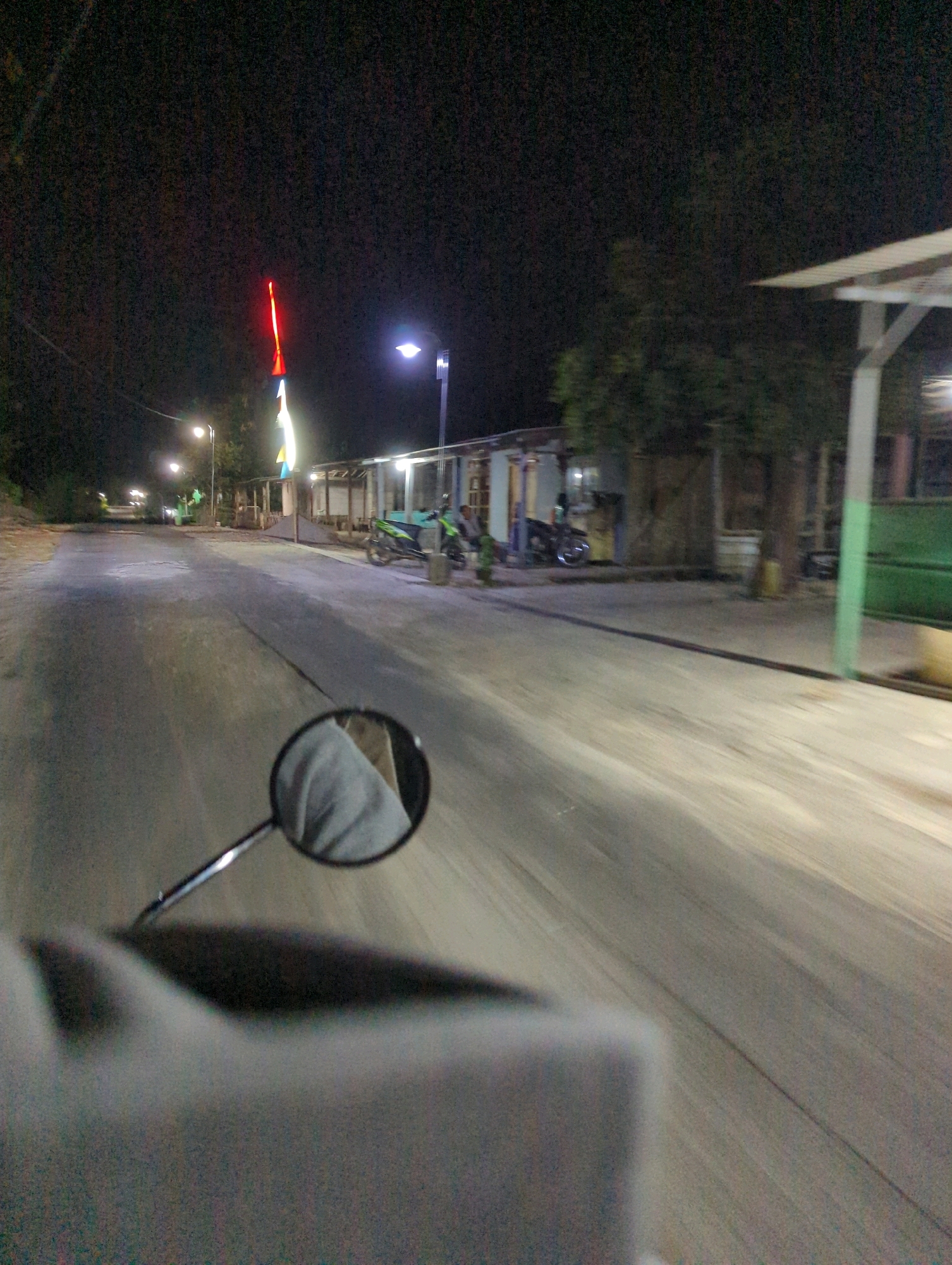 The image is a first-person perspective, taken at night, likely from a moving vehicle on a street in Senggrong, Indonesia. The road surface appears to be asphalt with a visible crack running down the center. On the right side of the road, there are several low-rise buildings with open fronts, some of which appear to be shops or residences. A green painted structure is also visible. Two motorcycles are parked in front of one of the buildings. In the distance, streetlights illuminate the road and the surrounding area, which includes trees and other vegetation. A prominent, tall, red and white illuminated structure, possibly a decoration or monument, stands out against the dark sky. A motorcycle side mirror in the foreground reflects a person's light-colored sleeve. The overall impression is a somewhat dimly lit, typical street scene at night in a rural or semi-urban Indonesian setting.