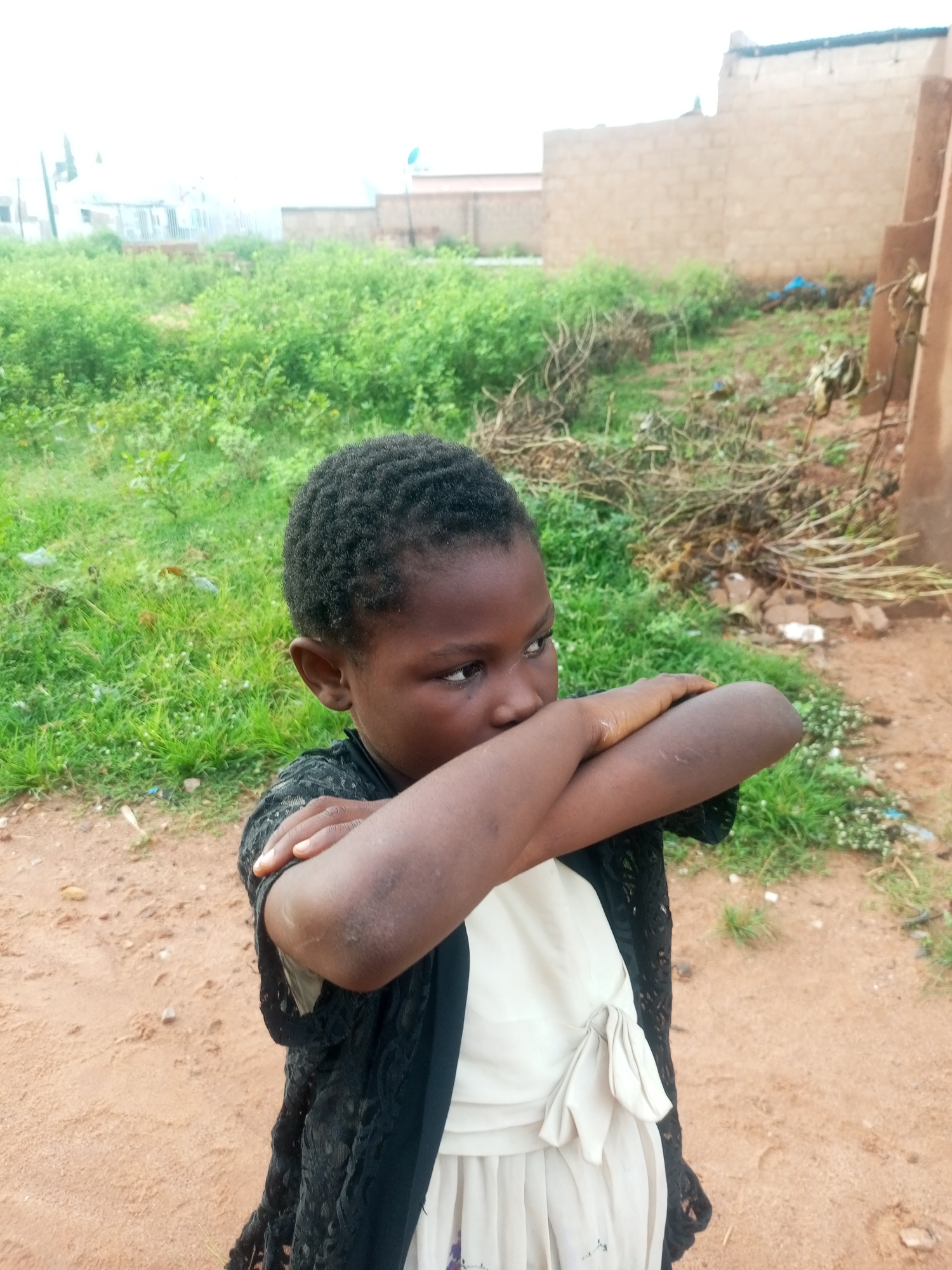A young Black girl, appearing to be of Nigerian descent, stands with her arms crossed in front of her chest, obscuring her mouth. She is wearing a white dress with a bow at the waist and a black lace overlay garment. The girl is positioned in a rural or undeveloped urban setting, with dry, sandy ground and sparse vegetation. In the background, there are unfinished brick buildings and overgrown bushes. The sky is overcast, suggesting it may be daytime in a tropical or sub-tropical region. The overall impression is one of a humble and somewhat neglected environment. The specific location in Tempure, Nigeria, is not visually discernible from the image itself.