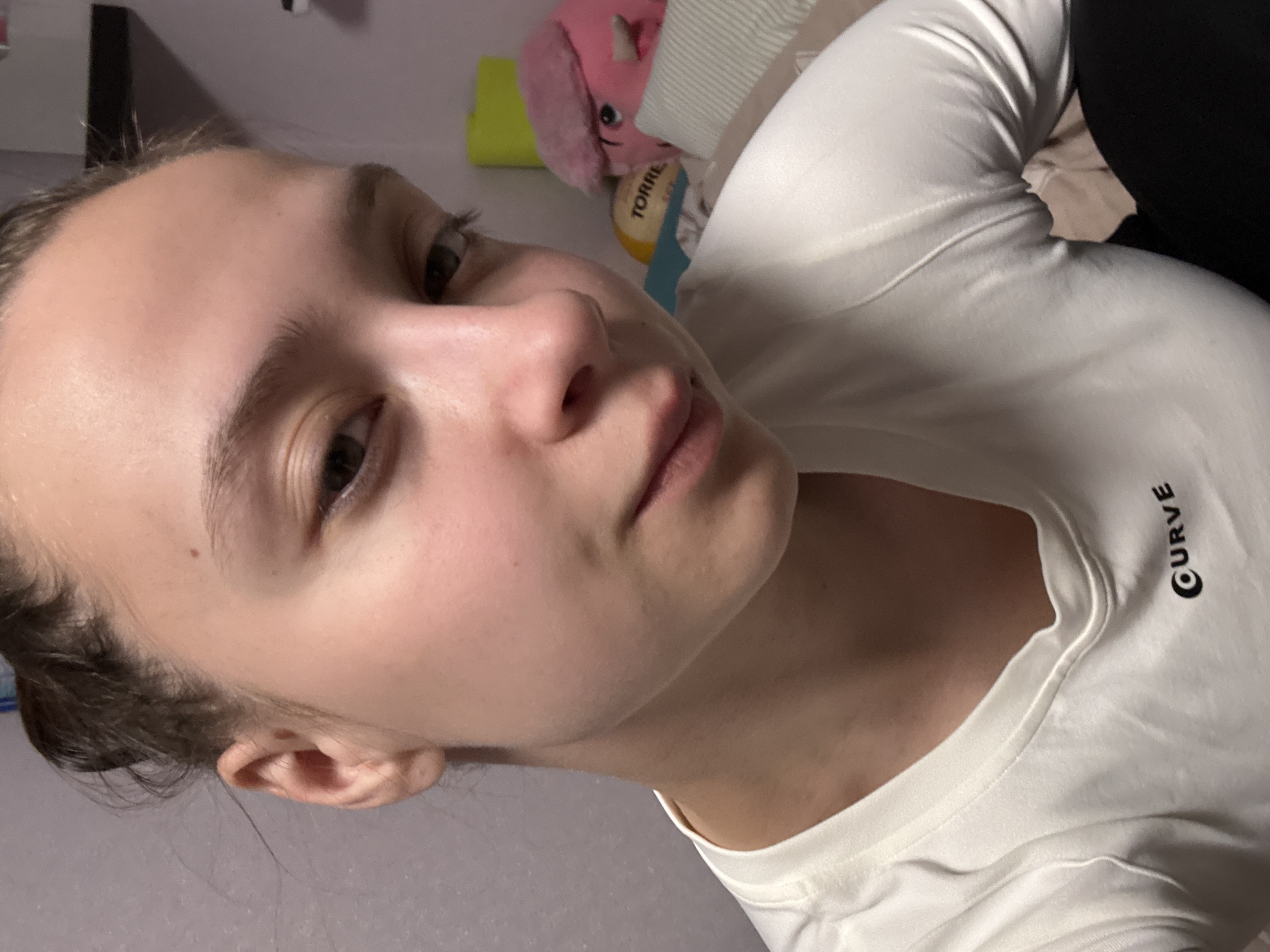 A close-up, high-angle photograph shows a young woman with fair skin, light brown hair pulled back, and light brown eyes. She is wearing a white long-sleeved shirt with the word "CURVE" printed on it. Her facial expression is neutral. In the blurred background, there are objects, including a pink stuffed animal, a yellow item, and a Torres package. The location is specified as Прикамский, Russia.