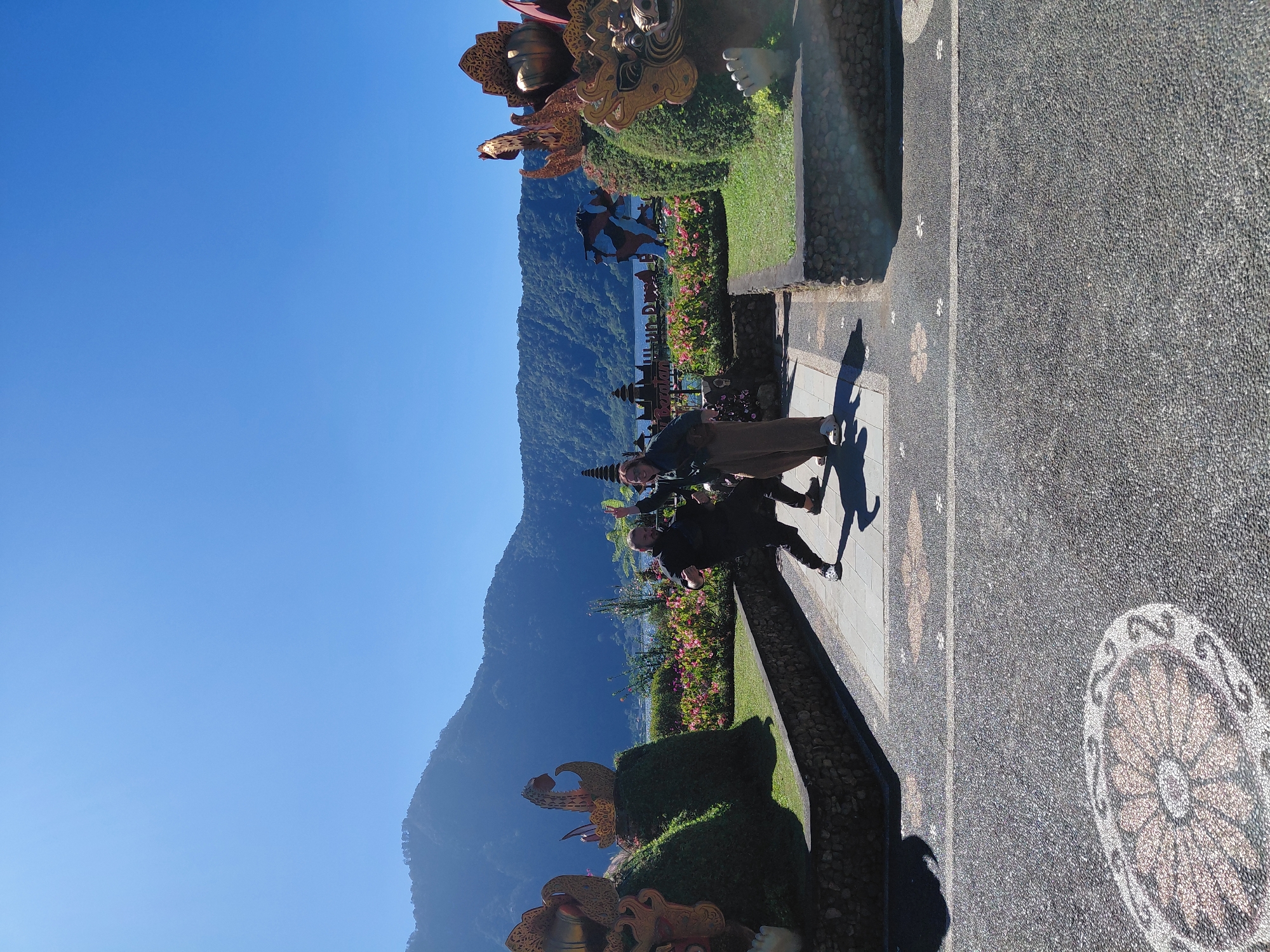 The image captures a vibrant outdoor scene at the Ulun Danu Bratan Temple complex, located in Baturiti, Indonesia. Two individuals are engaged in a playful, martial arts-like pose in the foreground, with one person in a black outfit and the other in a brown tunic and pants. Behind them, a lush garden of pink flowers creates a colorful backdrop, leading to a serene lake with mountains in the distance. Ornate golden sculptures of mythical creatures adorn the manicured hedges on either side of the pathway, adding a touch of grandeur to the location. The sky above is a clear, bright blue, indicating a sunny day. The ground is paved with a unique mosaic of small stones, featuring decorative floral patterns.