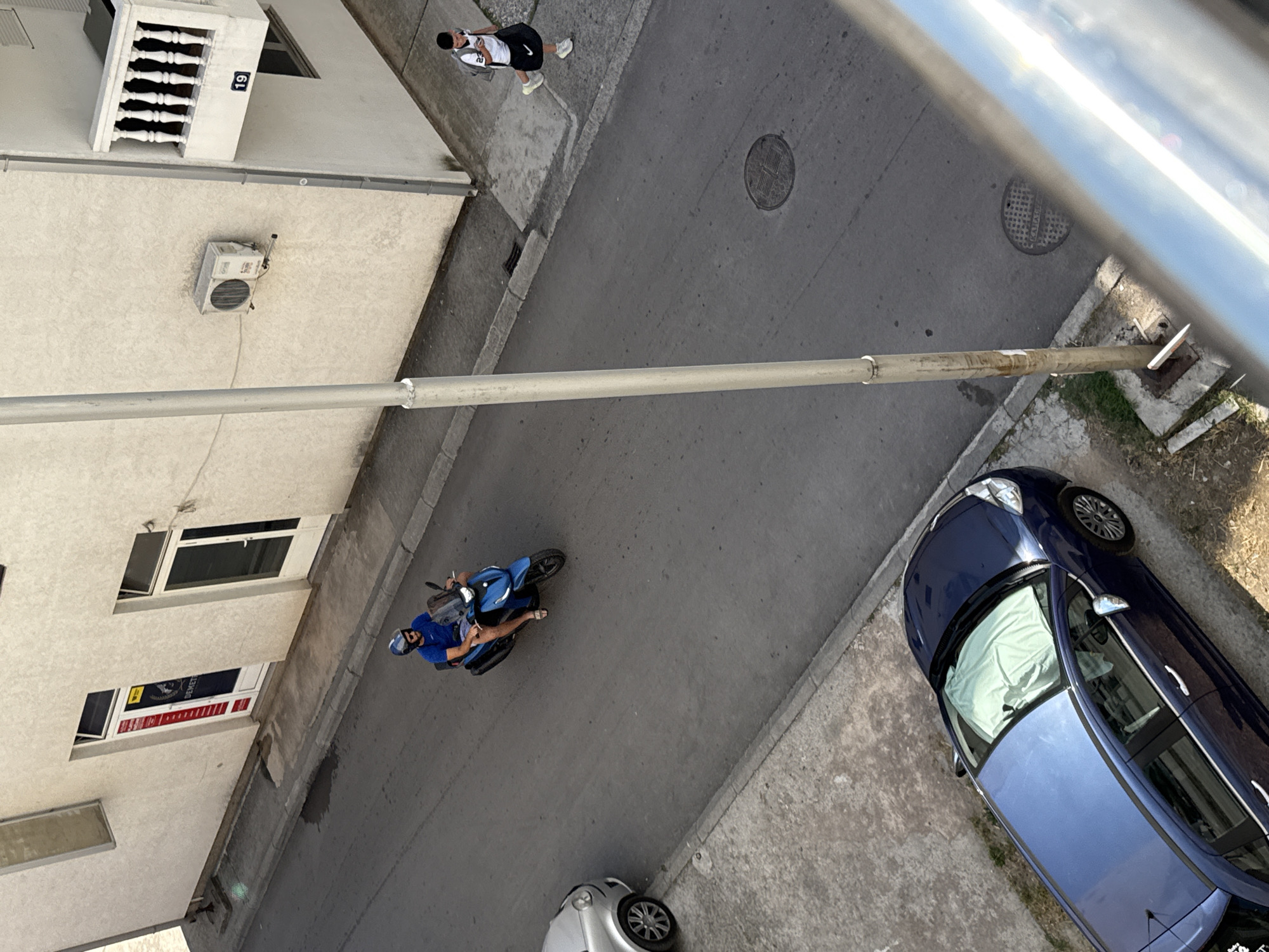 An overhead shot depicts a street scene in Budva, Montenegro. A person walks on the sidewalk near a building numbered "19". A man wearing a helmet and blue shirt rides a blue motorcycle on the street. A blue car is parked on the right side of the street, and a silver car is partially visible at the bottom of the frame. The buildings appear to be residential.