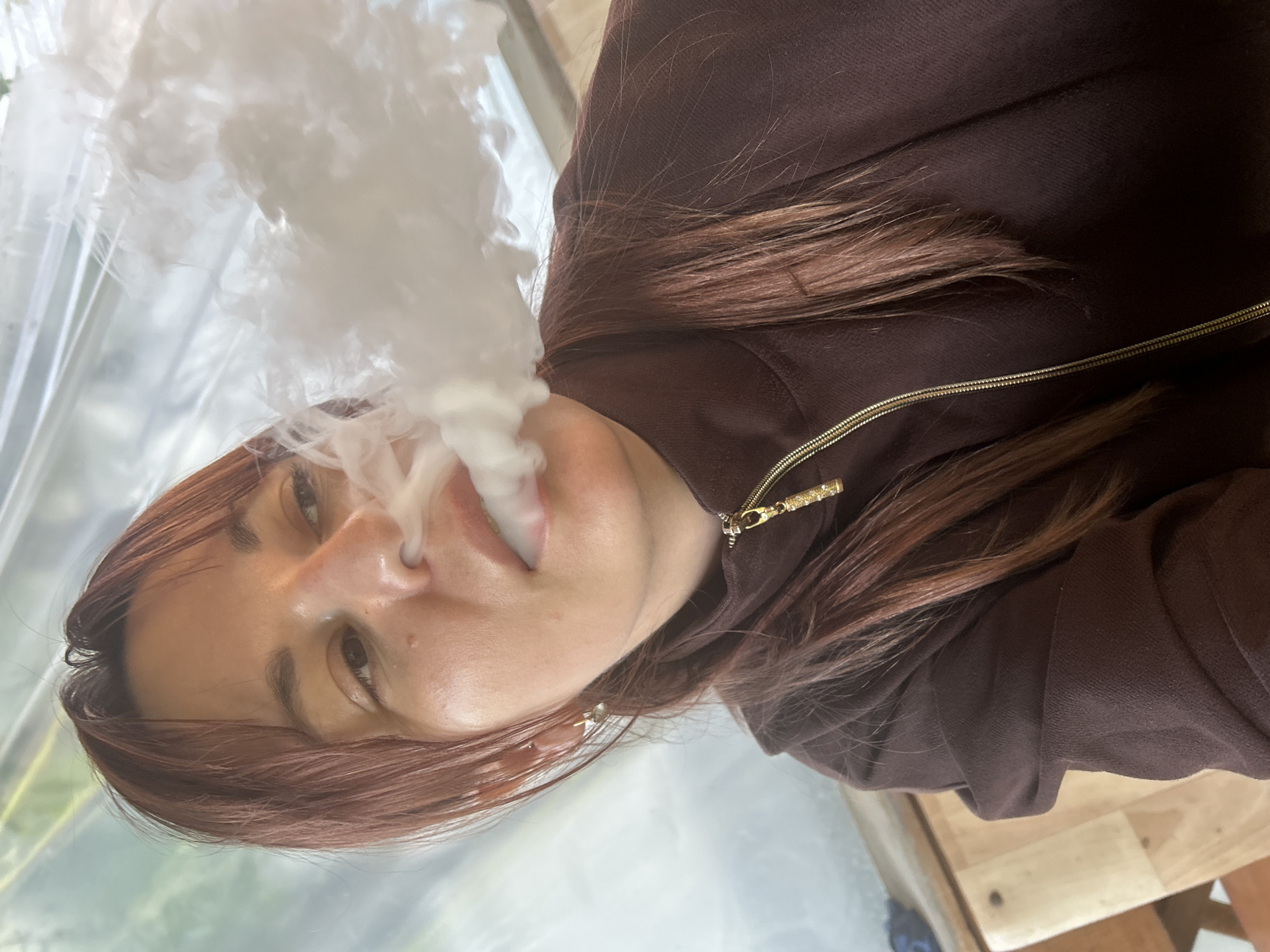 A woman with shoulder-length auburn hair, wearing a brown zip-up jacket with a gold zipper pull, exhales a cloud of white vapor, possibly from a vape or cigarette. The background is blurred but suggests an outdoor or sunlit setting, possibly a restaurant or patio. No evidence to confirm the location is Фоки, Russia.