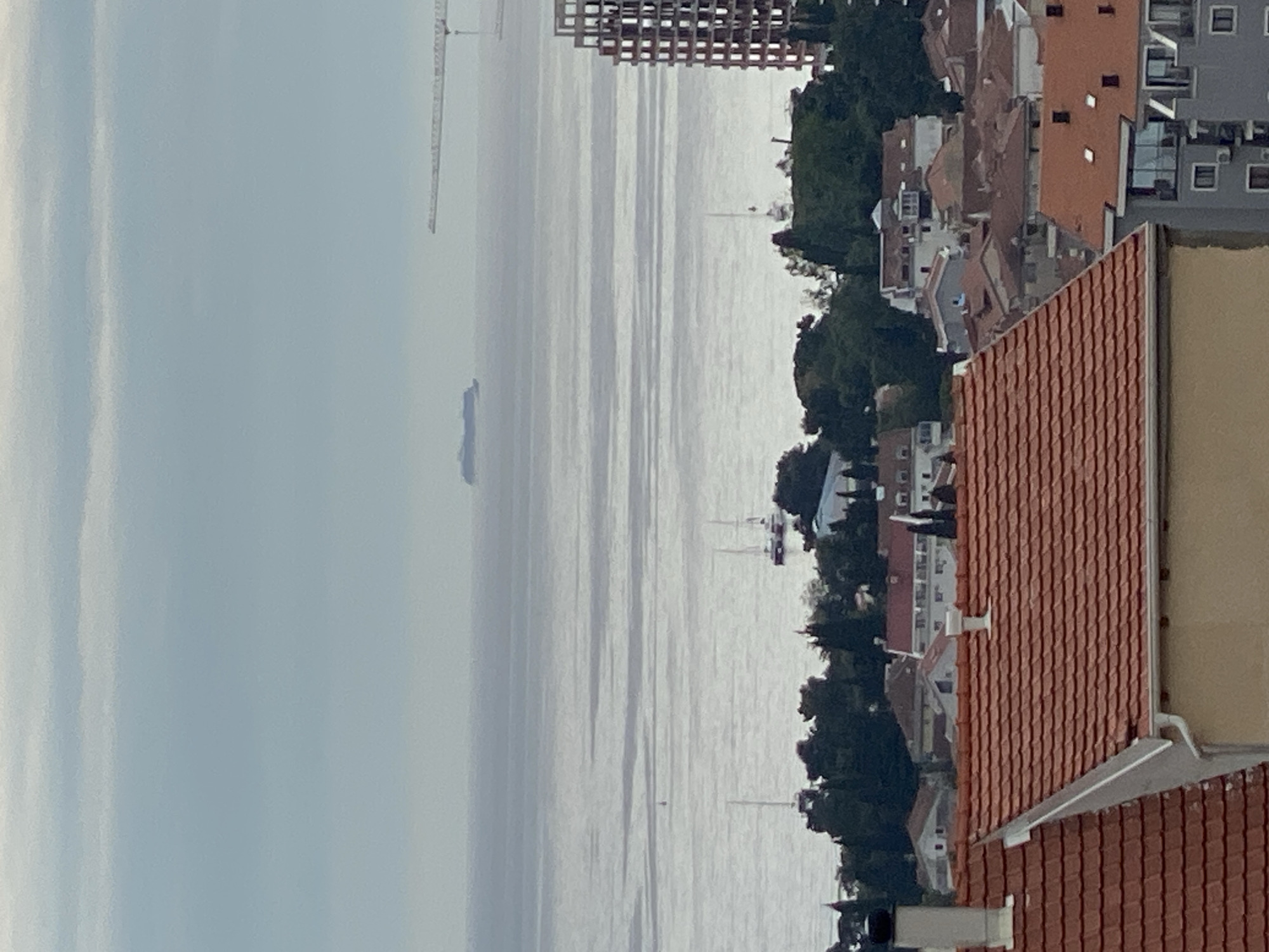 A horizontal, aerial photograph captures a coastal cityscape on a cloudy day. The foreground is dominated by rooftops, mostly covered in red terracotta tiles. Buildings vary in height and design, including some modern grey structures. The middle ground features a wide expanse of water, reflecting the overcast sky. A cargo ship and several sailboats are visible in the distance, along with a construction crane and an unfinished building. A strip of land with dense trees and scattered buildings separates the rooftops from the water. The image depicts the landscape in the city of Budva, Montenegro, overlooking the Adriatic Sea.