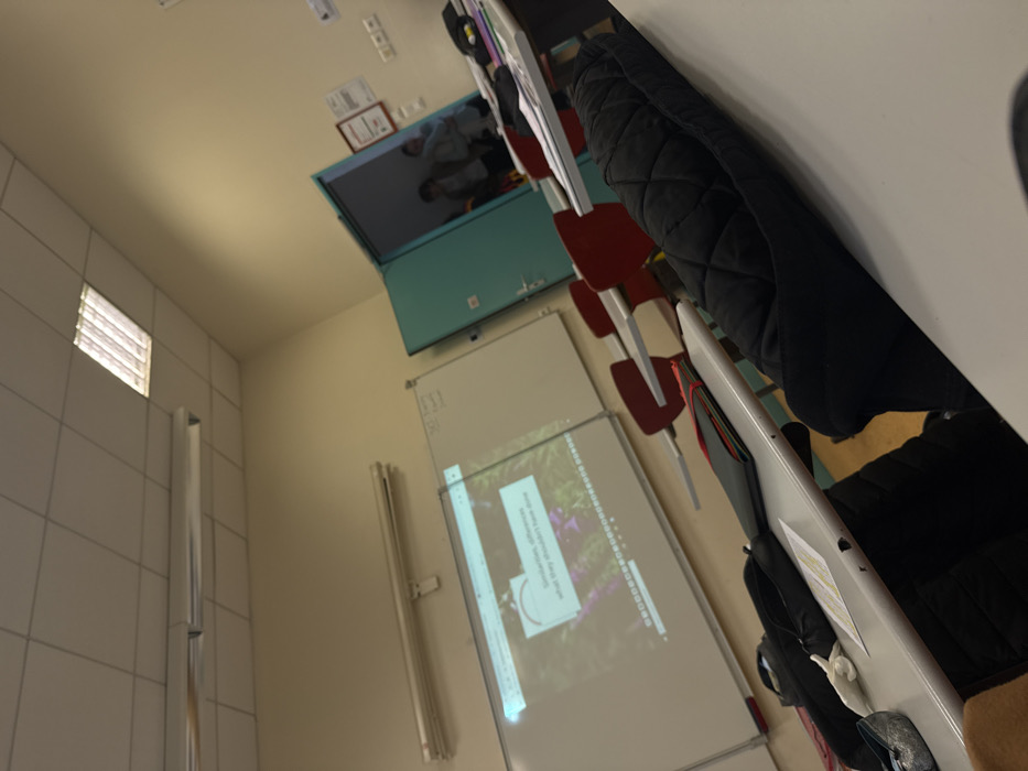 The image depicts the interior of a classroom or lecture hall in Albi, France, observed from a low angle at a desk. The room features cream-colored walls and a white acoustic tile drop ceiling with active fluorescent lighting fixtures. On the far wall, two whiteboards are visible. The left whiteboard displays a projected digital presentation featuring the text "Similarities, differences what they shouldn't have done" against a background image of purple flowers, with a red circular progress icon overlaid. The right whiteboard is largely blank.
Multiple light-colored student desks with attached red chairs are arranged in rows, receding into the background. A teal-colored door is partially open on the right side of the frame, revealing a narrow section of a hallway and three indistinct human figures standing or moving within it.
In the immediate foreground, on a desk to the right, rests a black quilted jacket or bag. On a desk to the left, a black bag, a pair of silver-colored ballet flats or similar shoes, a black binder/folder, and several papers (one yellow, one white) are present.