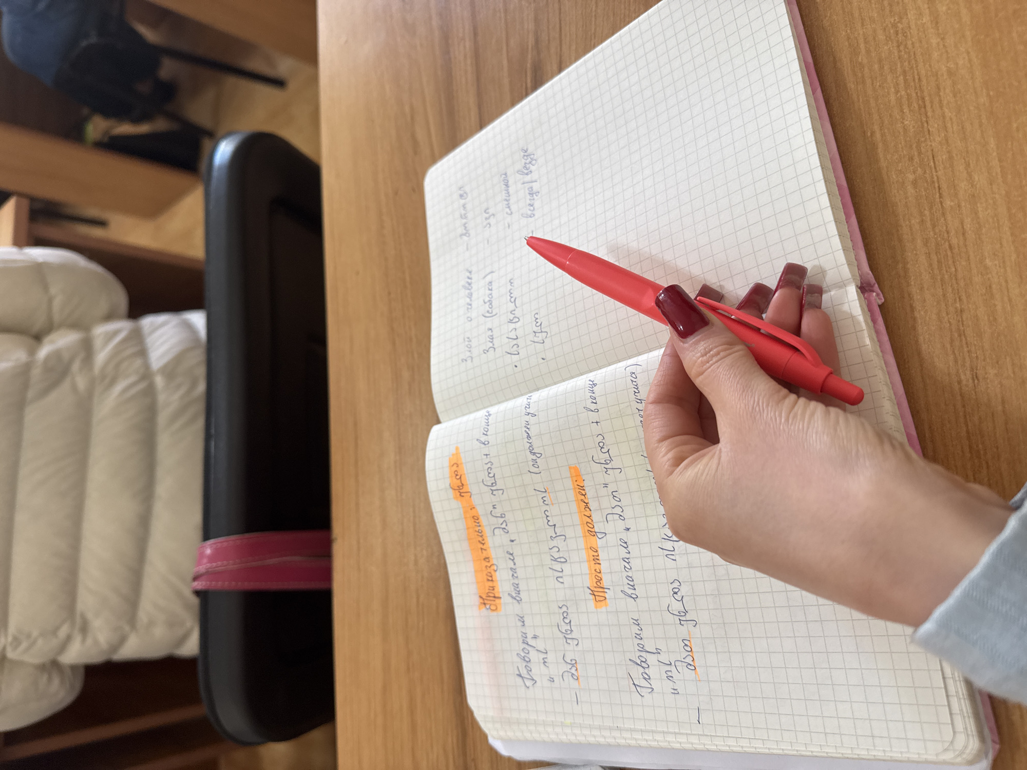 Here's a detailed description of the image:
**Scene:** The image captures a close-up view of an open notebook on a wooden desk. The notebook has a pink cover and graph paper pages, with handwriting in two languages (Georgian and Russian) and some highlighted sections in orange. A person's hand, with long, maroon-colored fingernails, holds a red pen poised over the open page, as if in the act of writing or underlining.
**Objects:**
* **Notebook:** Open, with text in two languages.
* **Pen:** Red, held in the hand.
* **Desk:** Wooden surface.
* **A black bag** or case is present on the left side.
* **A white blanket**
**Text content:**
The highlighted sections and written text seem to involve instructions, phrases, or vocabulary, possibly related to language learning or linguistics.
**Location:** It is hard to determine a specific location.