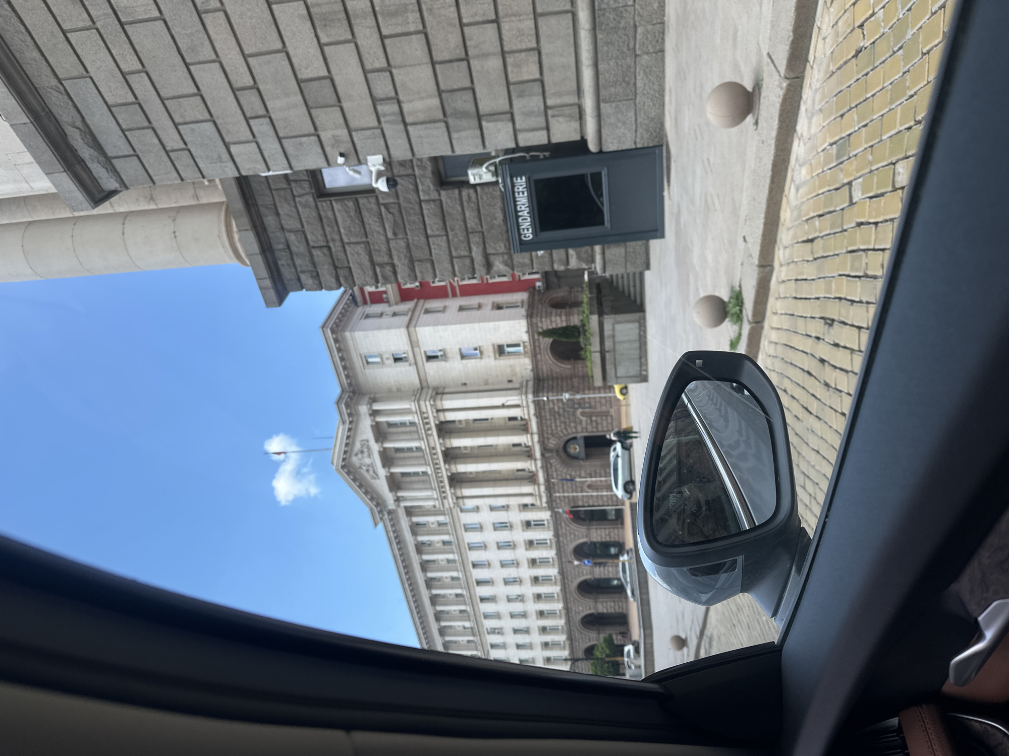 The image, captured from inside a vehicle looking out the passenger window, shows a street scene in Sofia, Bulgaria, under bright daylight. The photograph is oriented vertically, making the horizontal scene appear rotated.
In the foreground, elements of the car's interior are visible, including the dark window frame, a side mirror reflecting the street and buildings, and a glimpse of a tan seat.
The midground features a paved area with light-colored bricks leading up to a large building with a grey stone facade. Prominently positioned against this building is a black security booth with the word "GENDARMERIE" written in white capital letters. Several security cameras are mounted on the building's facade above and around this booth.
In the background, a grand, neoclassical-style building with numerous windows, white columns, and architectural details dominates the view. The Cyrillic text "МИНИСТЕРСКИ СЪВЕТ" (Ministerski Sŭvet), which translates to "Council of Ministers," is clearly visible on its facade, identifying it as a significant government building in Sofia. A smaller building with red and white sections is partially visible behind the stone facade. The sky is clear and bright blue with a few scattered white clouds, indicating sunny weather. A flag on a flagpole is seen against the sky. Several cars are parked or visible along the street near the main building.
The scene portrays a calm daytime moment, likely from a vehicle stopped or moving slowly past a prominent administrative area in the city.