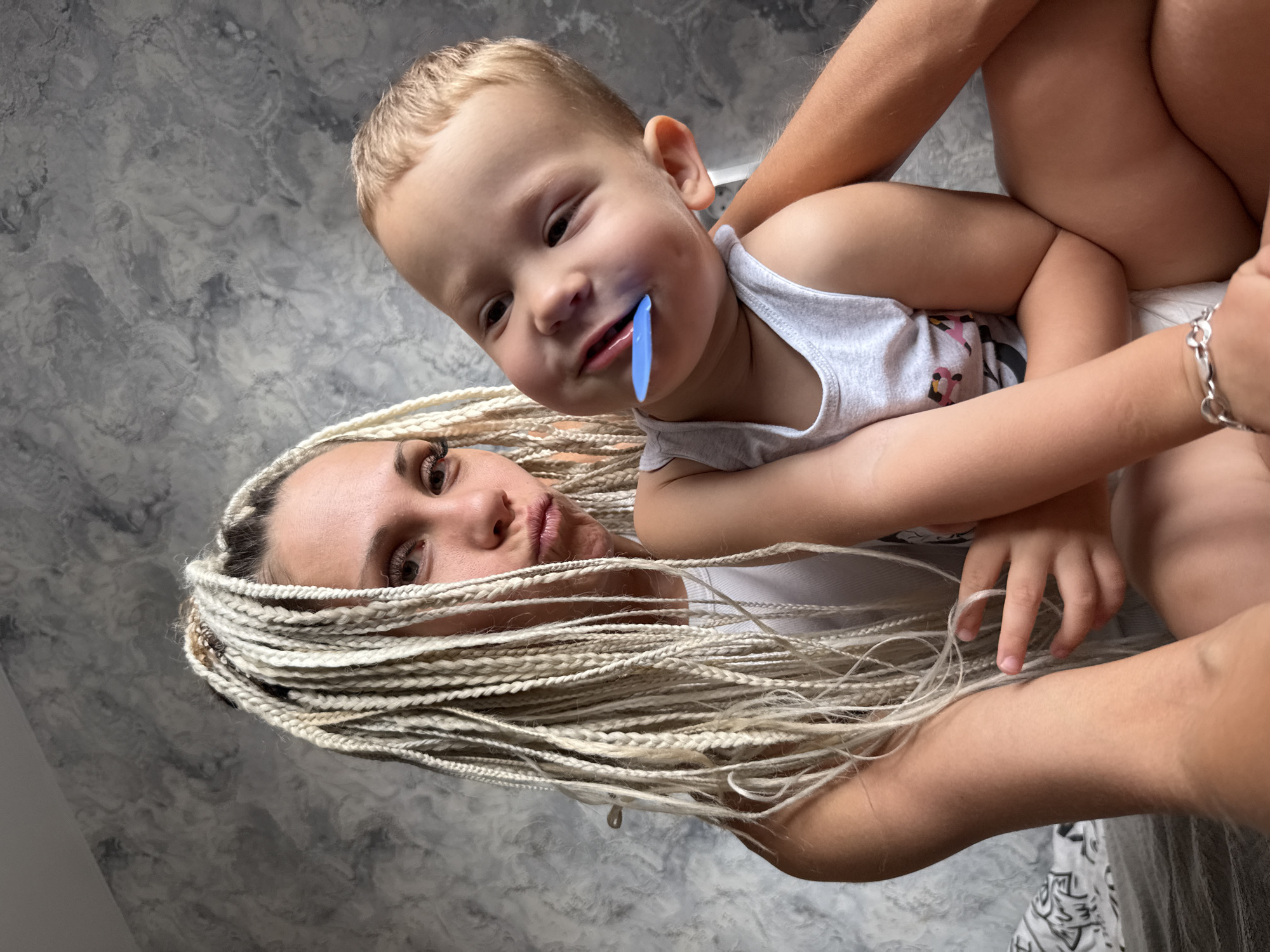 This is a close-up, high-angle, outdoor selfie of a woman and a young child, likely mother and son, in Rusanovo, Russia. The woman, with blonde braided hair that covers part of her face, is looking towards the camera with an amused expression. Her eyes are dark brown and she is wearing a white top. The child, with fair hair, is held by the woman and is looking at the camera with a playful expression. A small blue object is in his mouth. He is wearing a white tank top. The background is a textured grey wall, possibly inside a home. The lighting suggests it is daytime. The overall mood of the image is affectionate and lighthearted. There is no visible text in the image.
