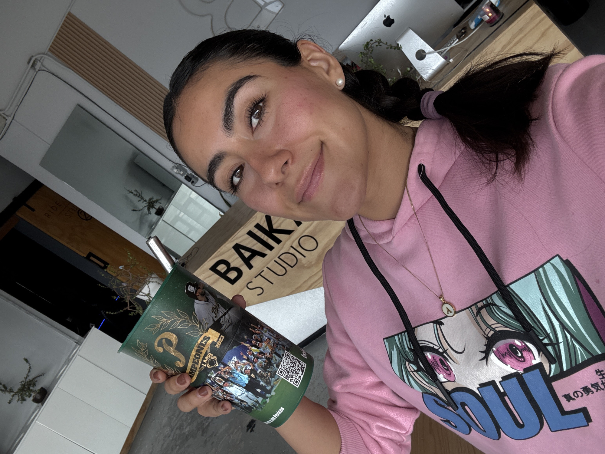 A woman is holding a mate cup with the words "CAMPEONES LMB 2023" and a team photo, indicating the Puebla Pericos baseball team won the Mexican Baseball League (LMB) championship in 2023. The cup also has a QR code. She is wearing a pink hoodie with an anime print. Behind her is a sign that says "BAIK STUDIO." The setting is likely indoors, possibly in Puebla, Mexico, given the baseball team affiliation. A ponytail can be seen on the upper right of the image.