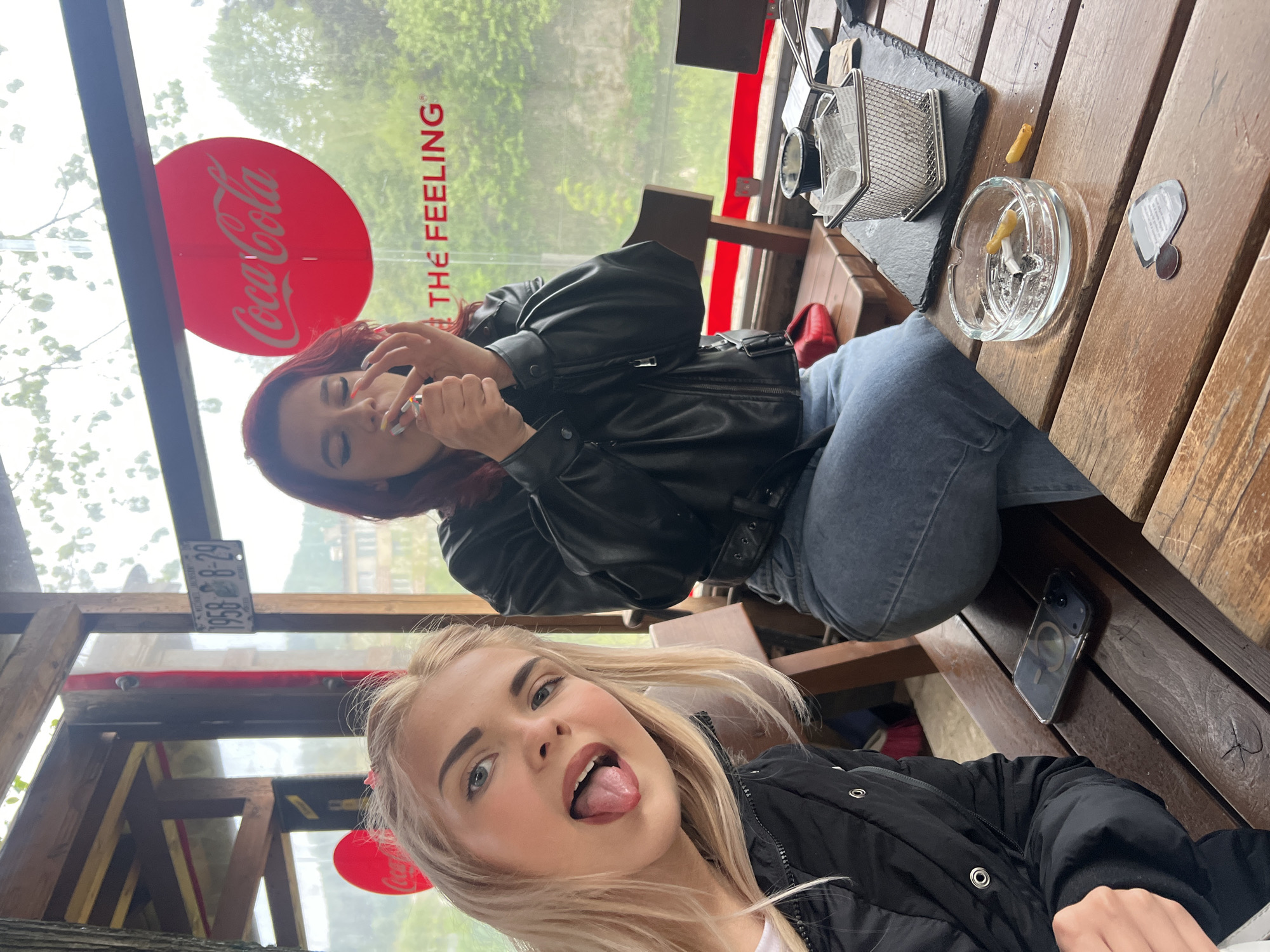 The image captures two women at a table within what appears to be a cafe or restaurant. The woman in the foreground has blonde hair and is sticking out her tongue in a playful manner, wearing a black jacket. The woman in the background has red hair and is wearing a black leather jacket and blue jeans, holding a lit cigarette. The setting appears to be a place in city of Unknown, Unknown, identifiable by a license plate reading "1958 B-29". The table holds an ashtray with cigarette butts, a wire basket, a menu or card, and a cell phone. A red "Coca-Cola THE FEELING" sign is visible on the window behind the women.
